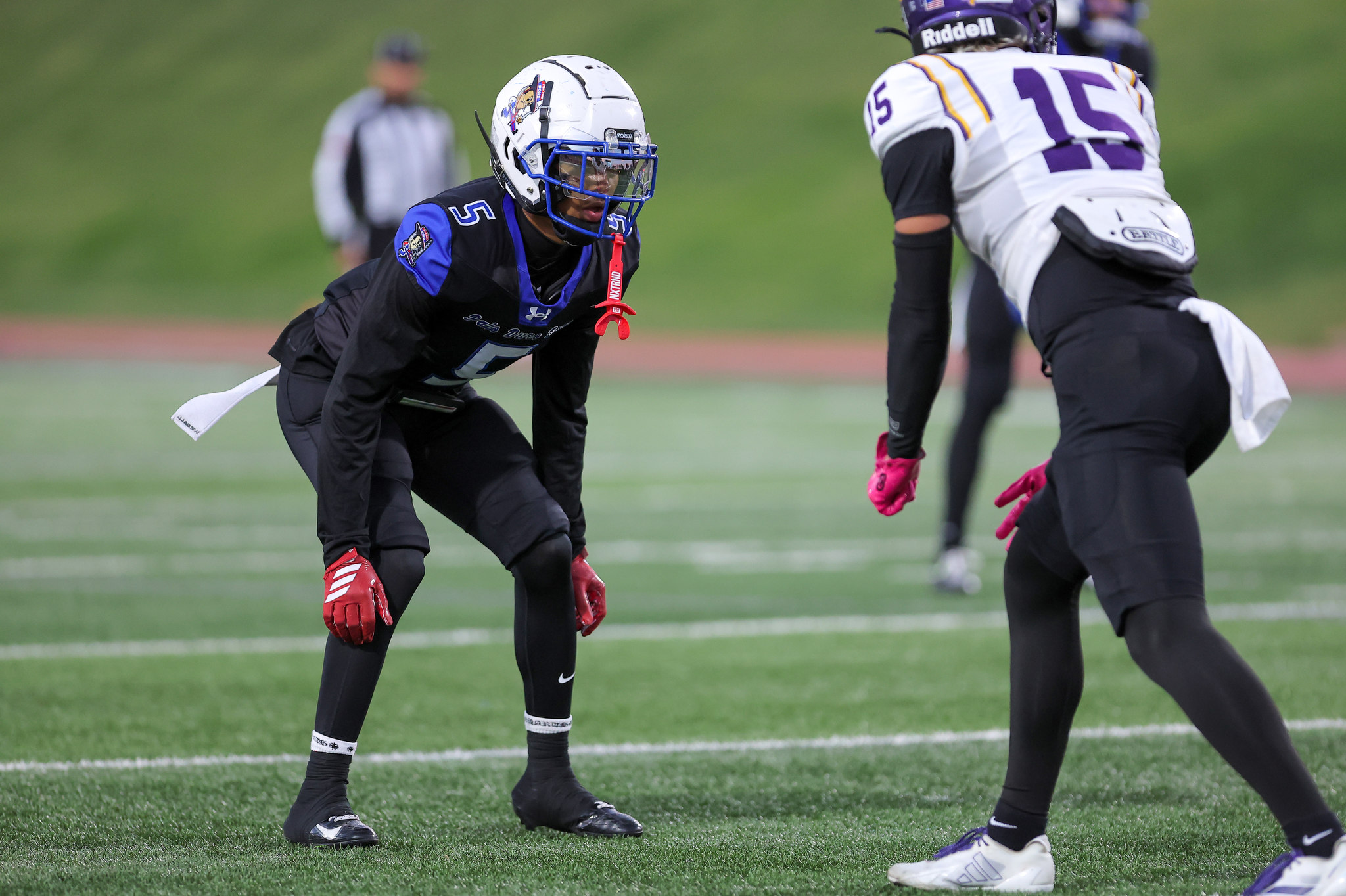 Two football players face off in a drill on a green field; one in a dark uniform with a white helmet, the other in a white jersey with purple numbers.