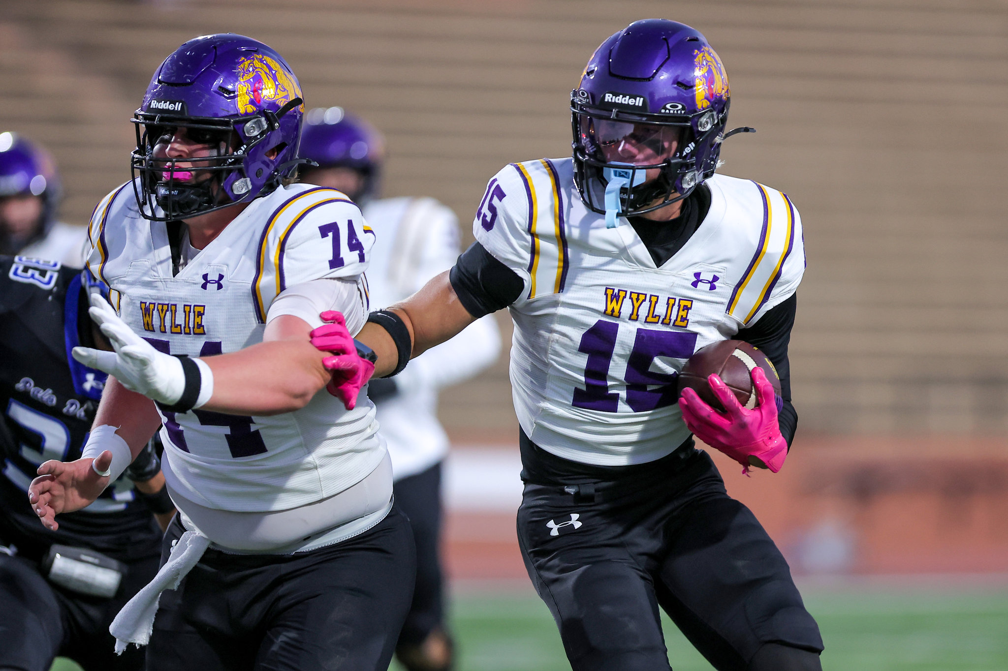 Two football players from the Wylie team in white jerseys with purple and gold trim, one carrying a ball as they run side by side in a game.