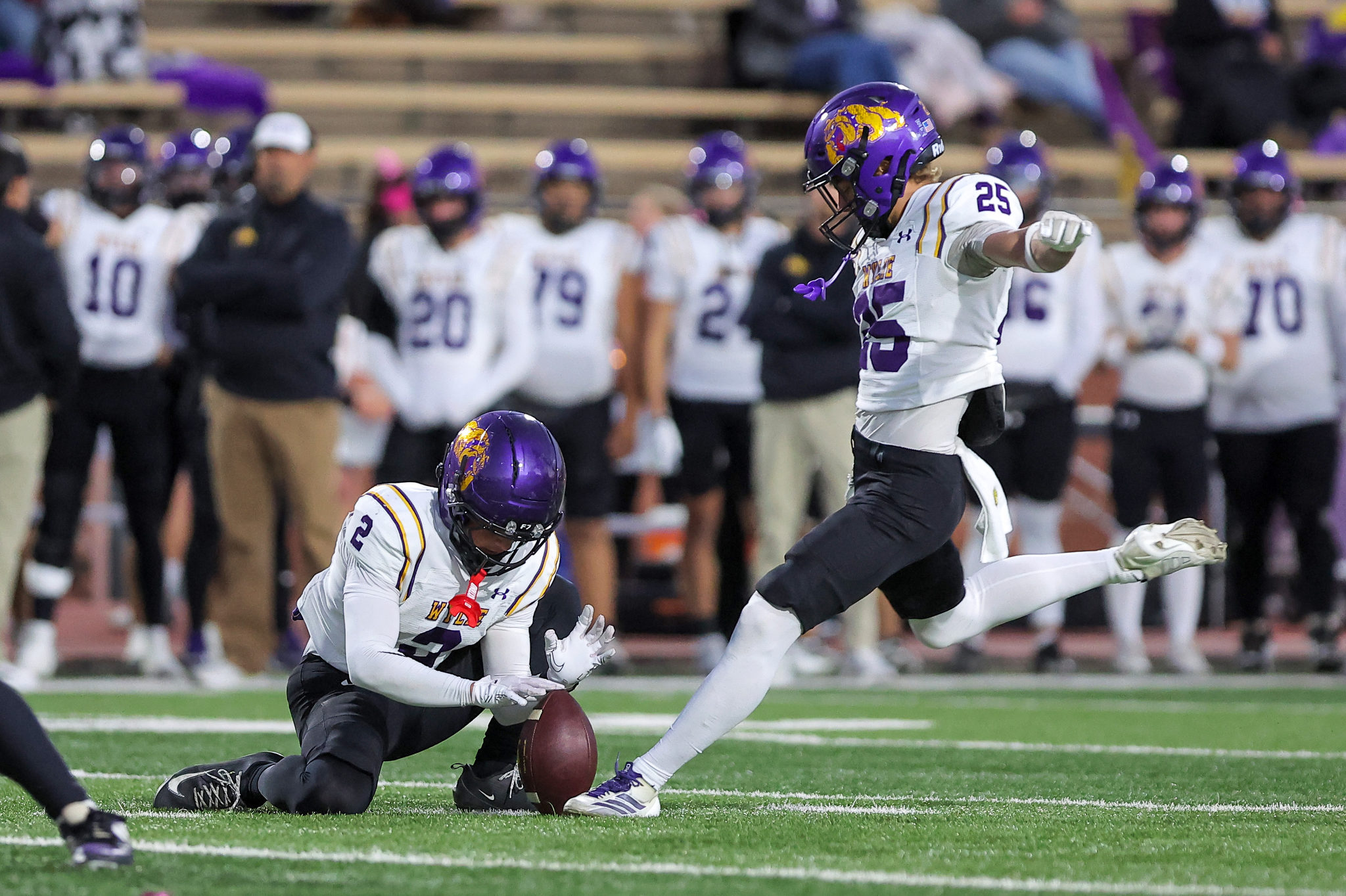 Football players in white and purple uniforms reach for a loose ball on a green field as a teammate dives nearby.
