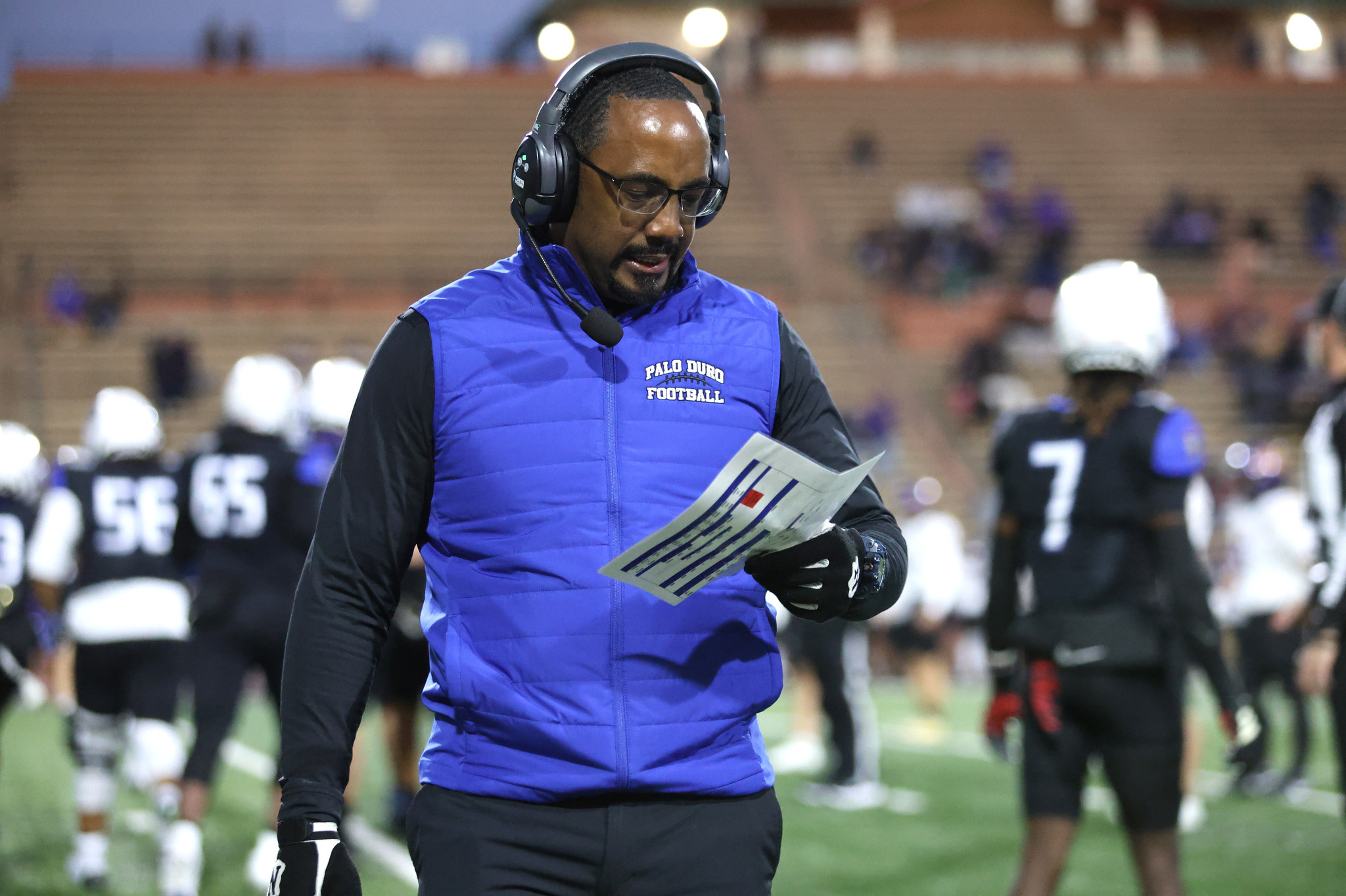 Football coach wearing a blue vest and headset reviews plays on the sideline with a clipboard in hand.