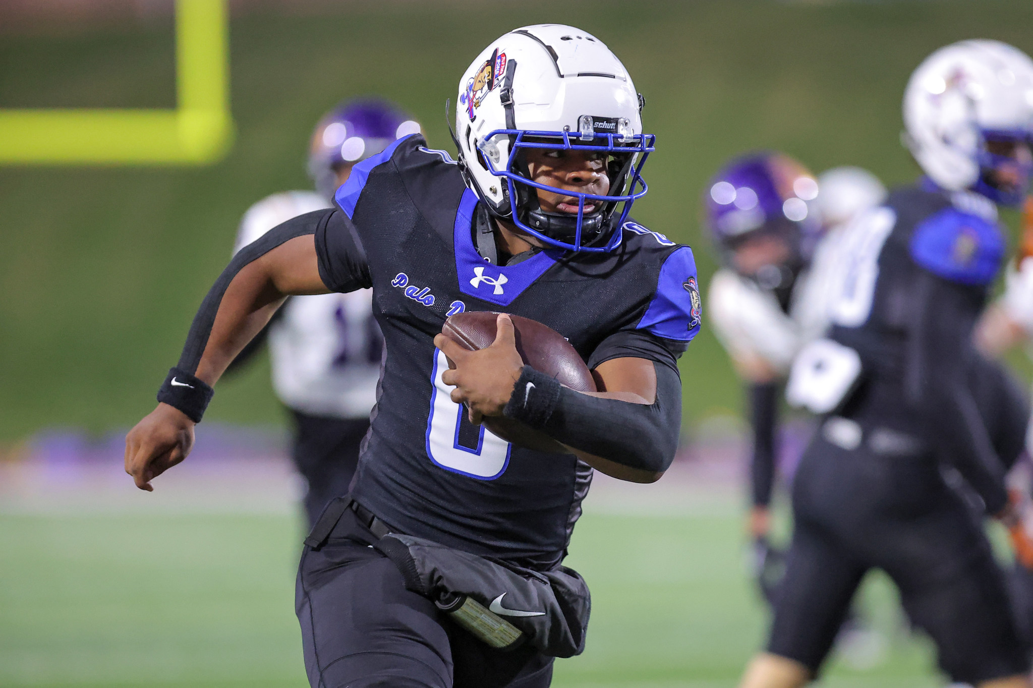 Football player in black and purple jersey runs with the ball during a game, helmet and pads visible, field in the background