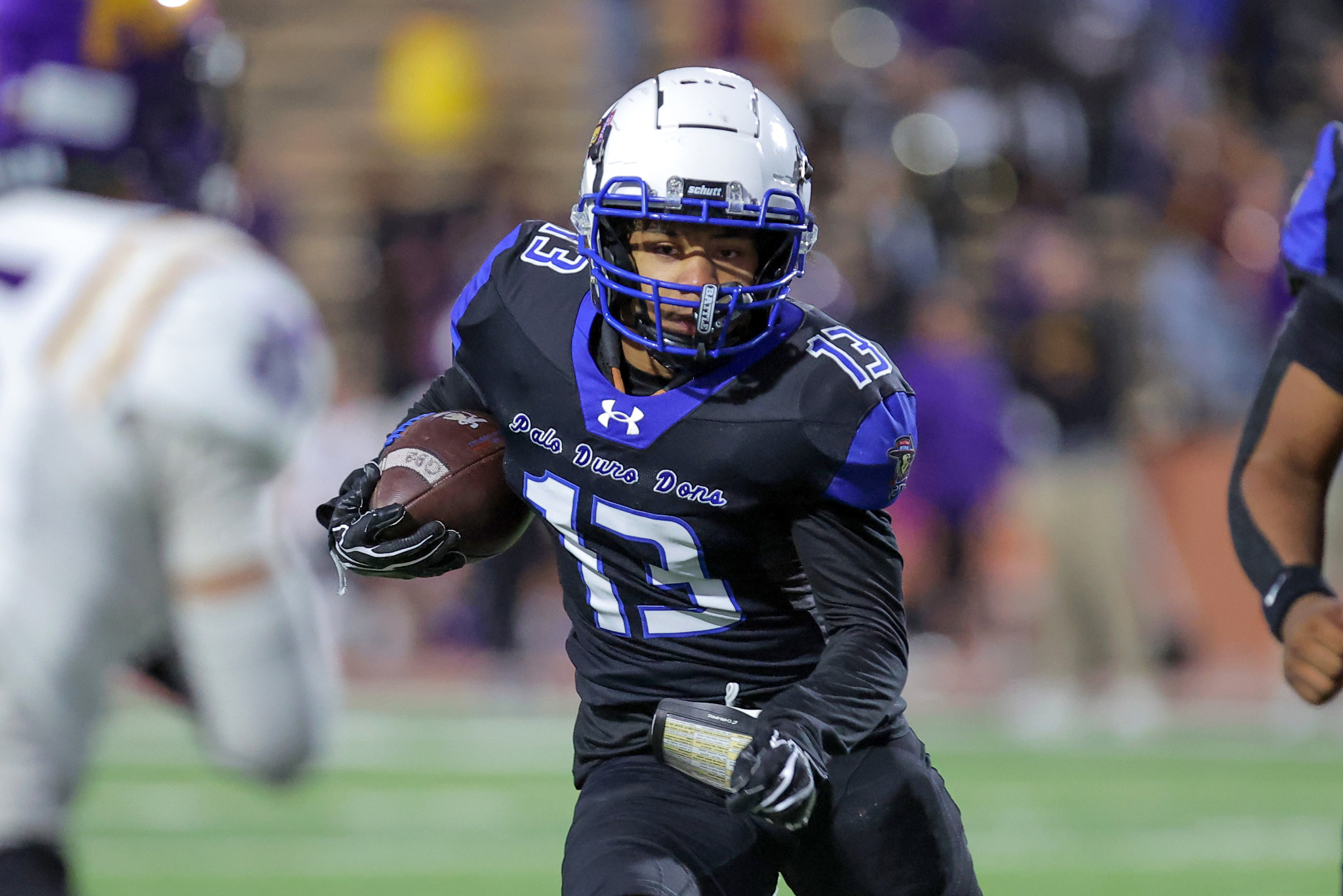 Football player in black and blue uniform runs with ball toward camera on a field, white helmet and blue facemask visible