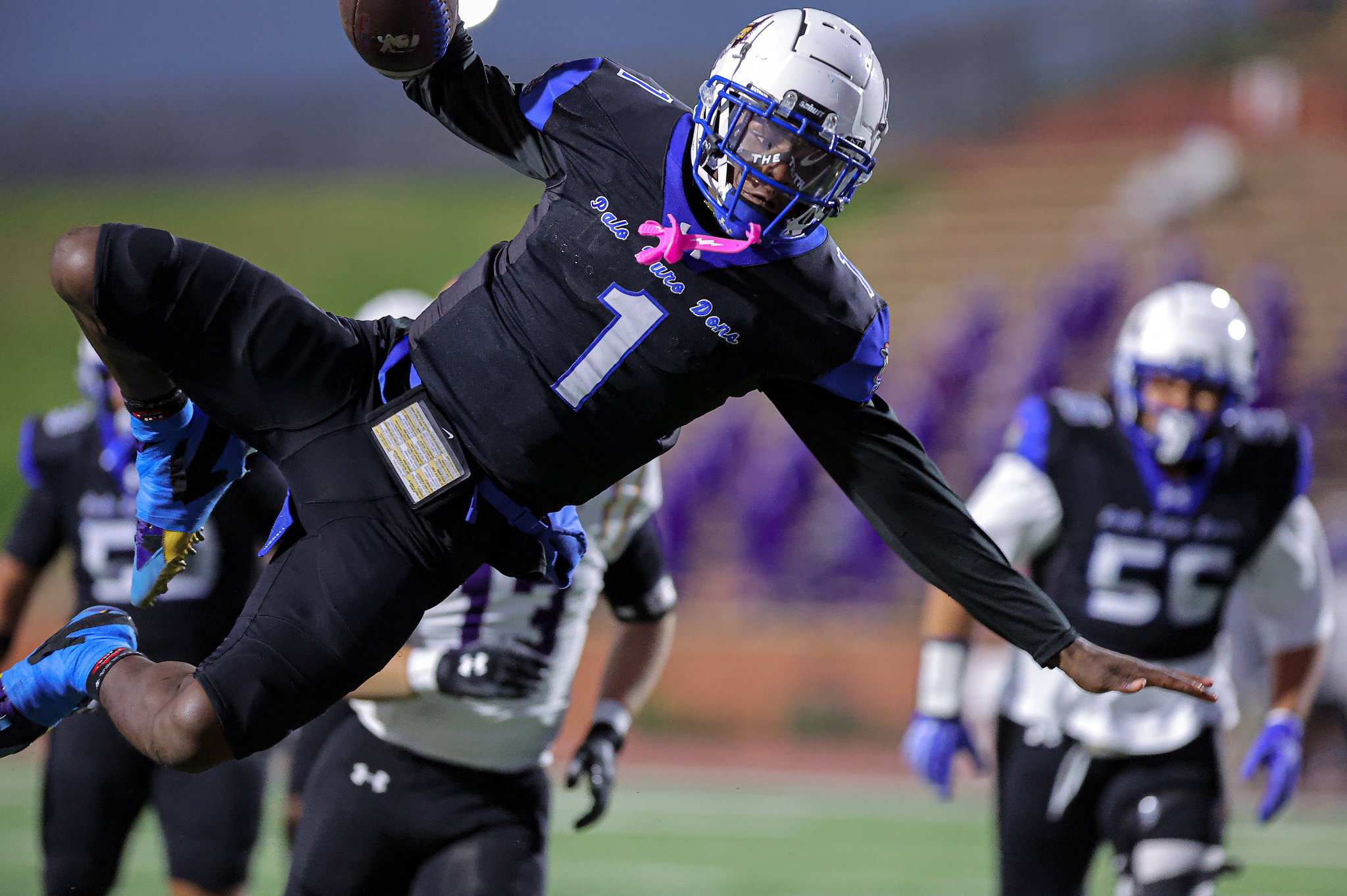 Football players in black and blue uniforms collide mid‑play on a field; one leaping with arms extended toward the ball under a blue sky/stadium backdrop.