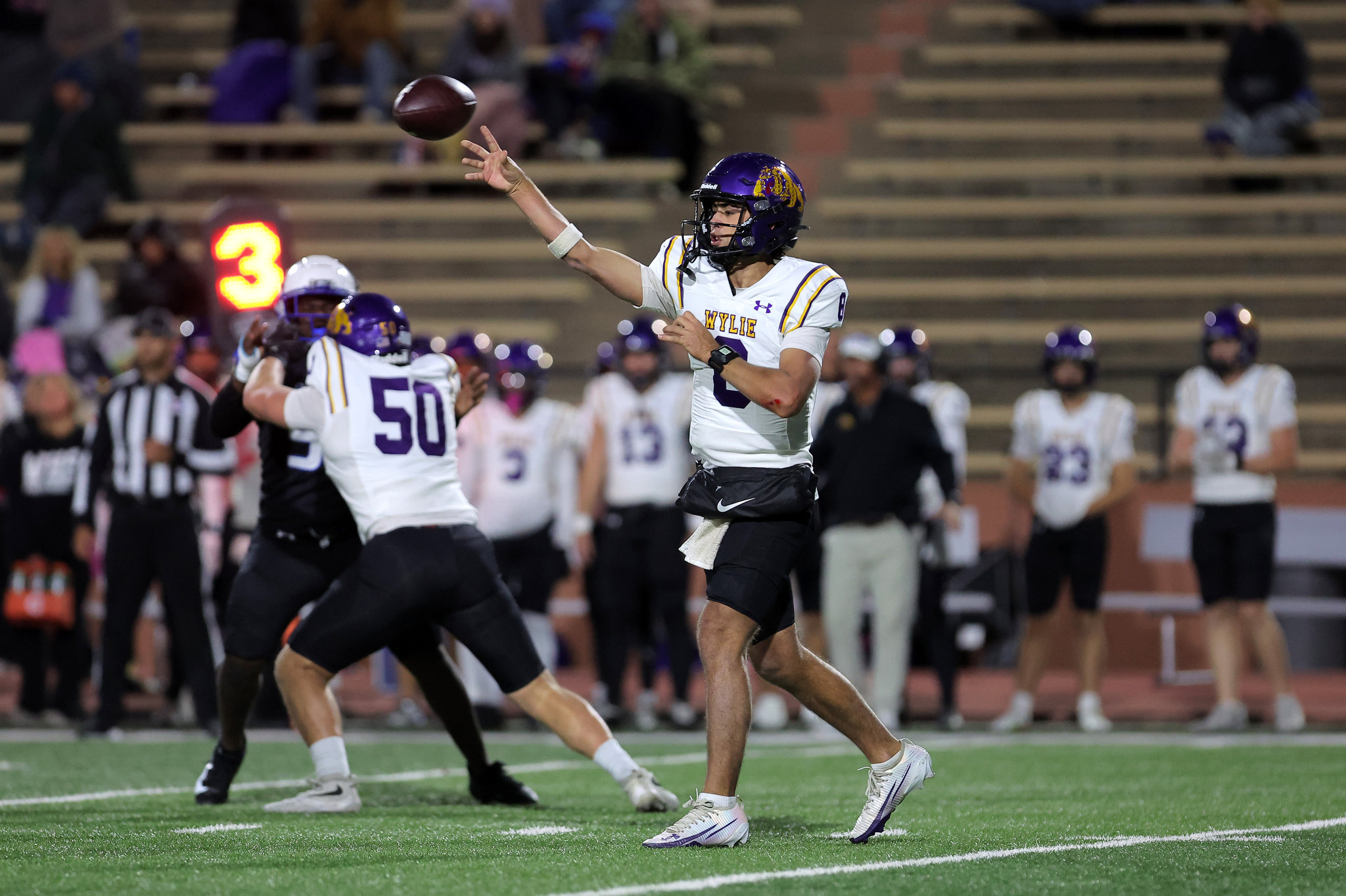 Quarterback in a white and purple uniform throws a football during a high school game on a green turf field.