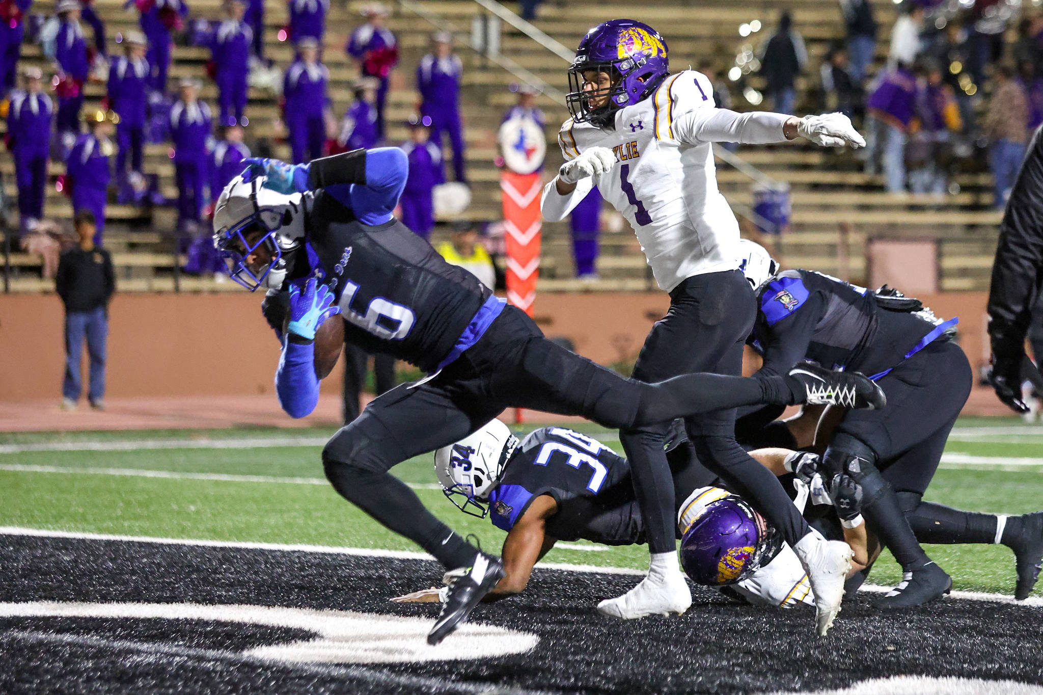 Football players in white and purple uniforms collide on a football field during a nighttime game; a white-uniformed runner (#1) is tackled as others dive and fall around him with a purple-clad defender in the foreground.