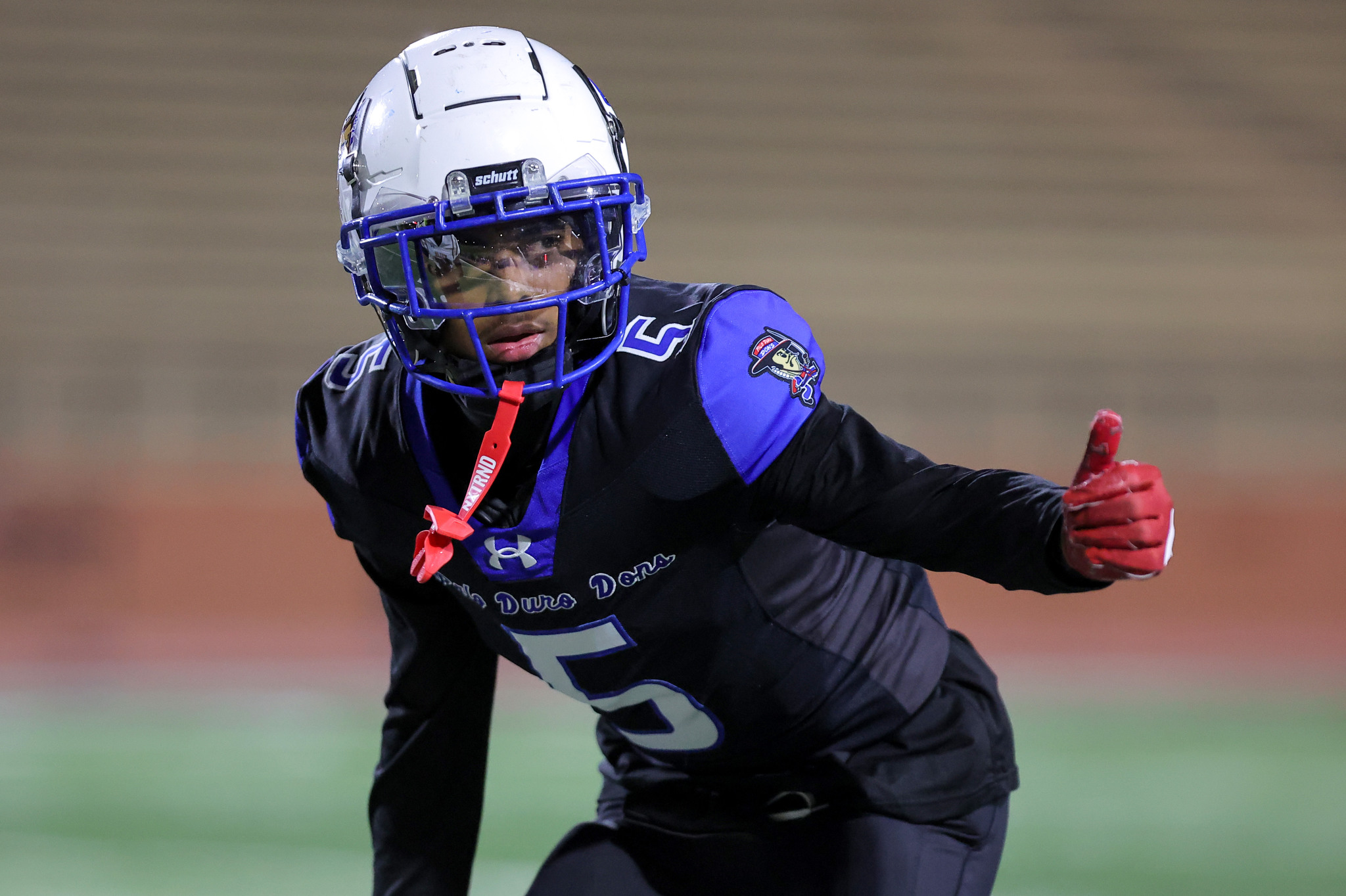 American football player in a blue-and-black uniform extends an arm during a game, wearing a white helmet with a blue face guard and red gloves on a green field