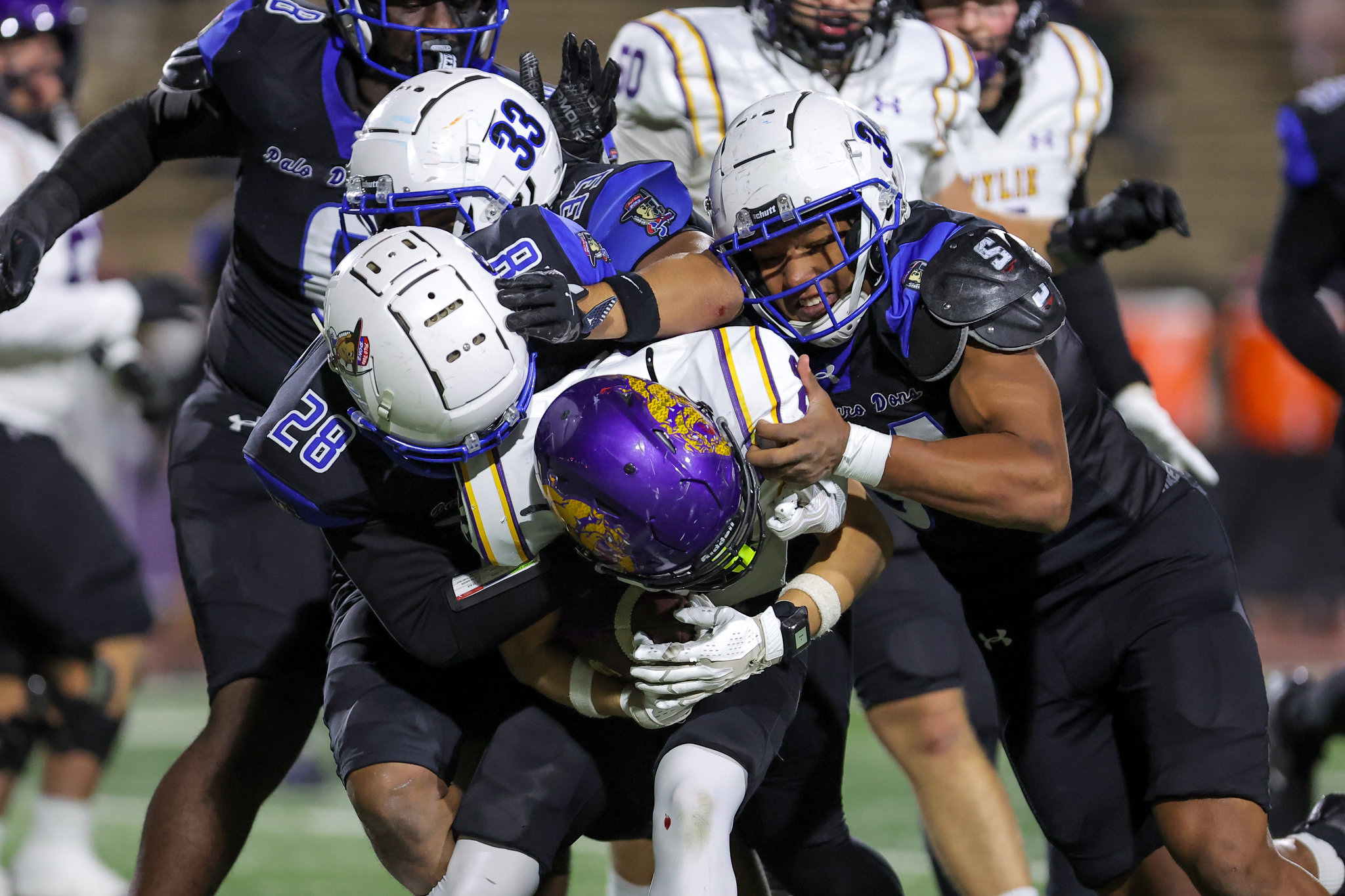 Multiple football players in blue, white, and black uniforms pile onto a tackled player wearing a purple and yellow helmet on a green field.