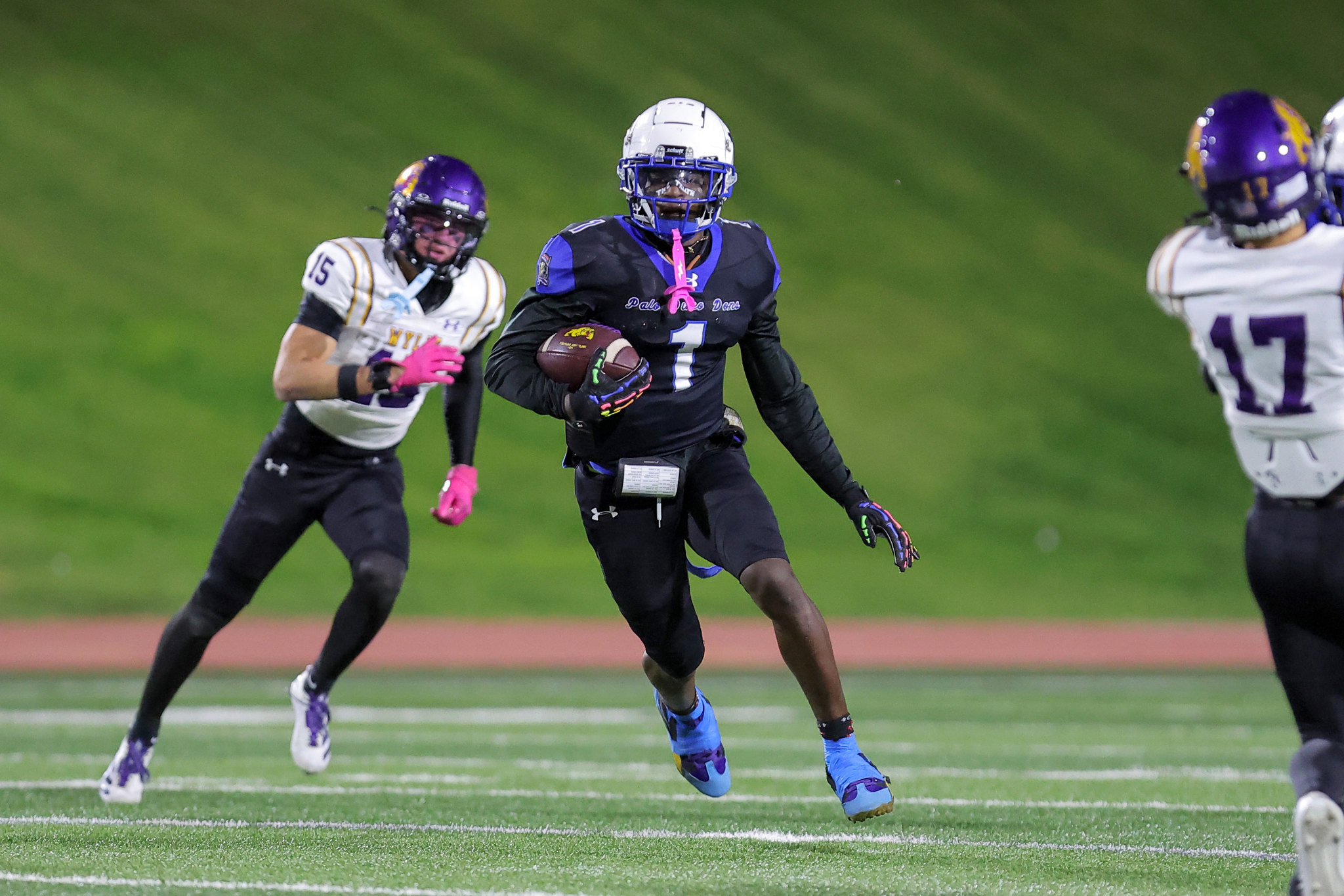 A football player in a black uniform with blue trim runs with the ball on a field, defenders in white uniforms nearby, night game lighting.