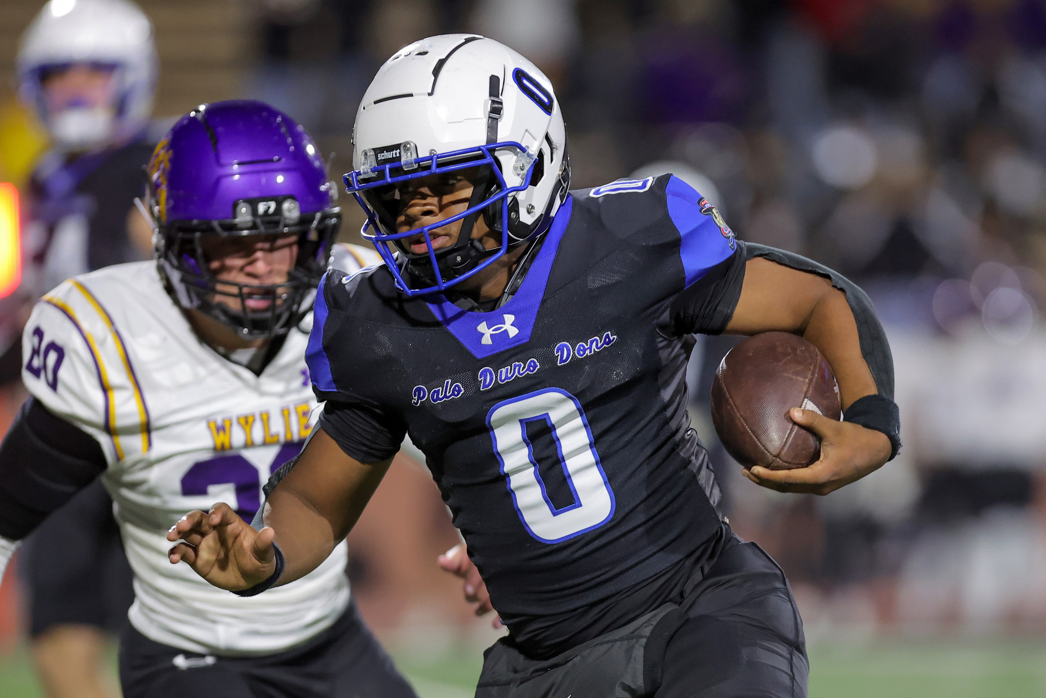 Football player in blue and black uniform runs with the ball during a game, gripping it securely as defenders close in.
