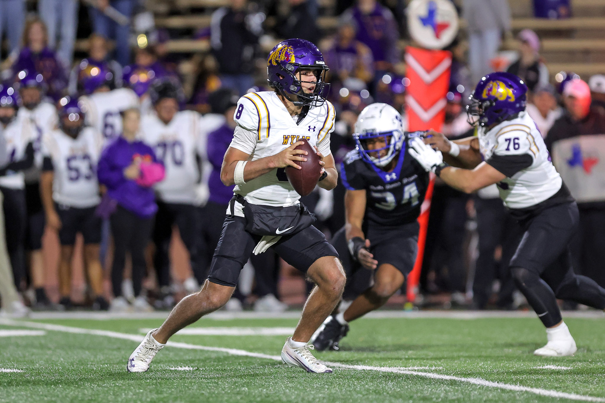 Football quarterback in a white and purple uniform runs with the ball as defenders pursue him on a green turf field; crowd in purple jerseys in the background.
