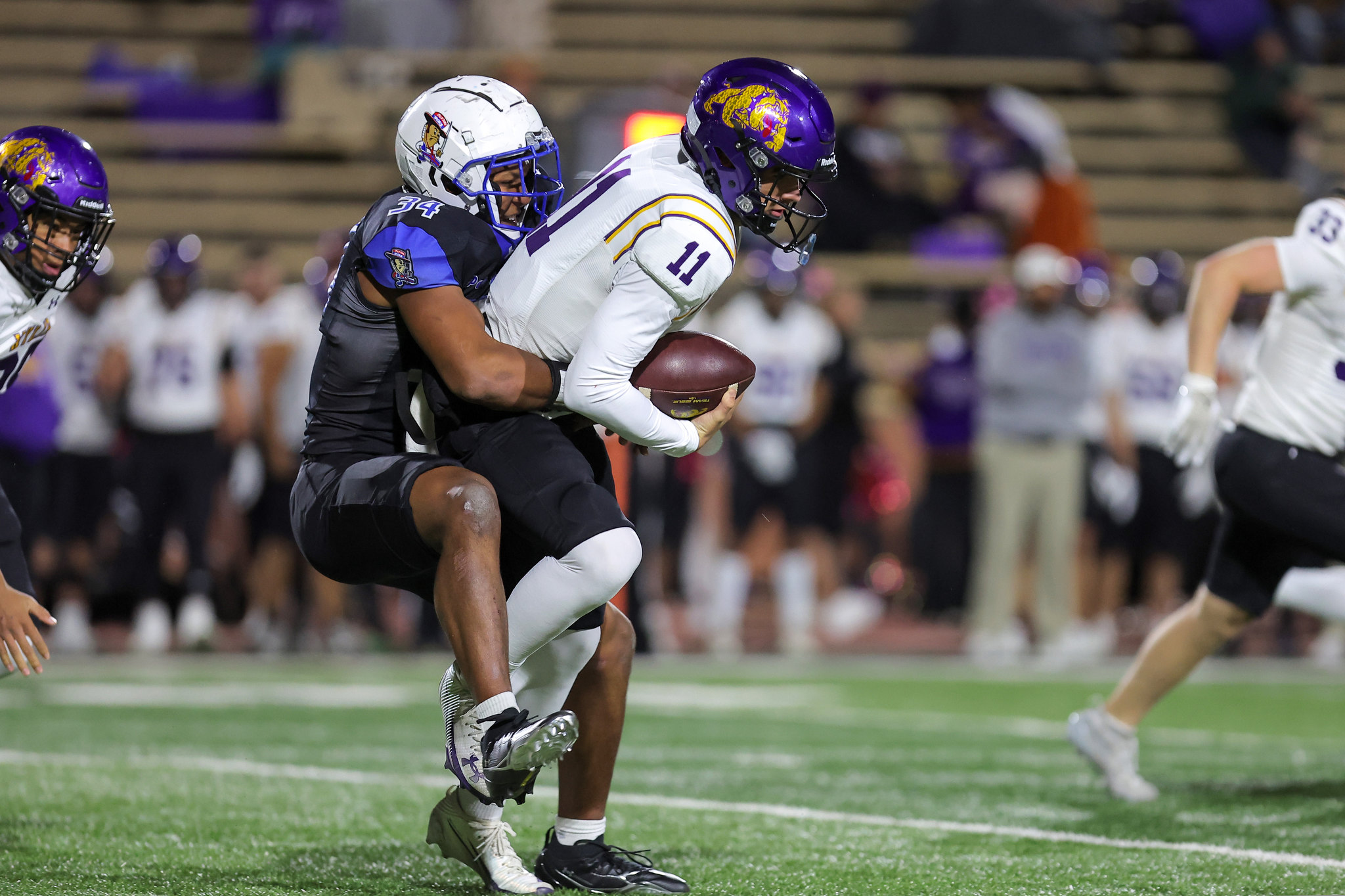 Football player in white jersey (number 11) carries the ball as a defender in a purple helmet tackles him from behind on a green field.