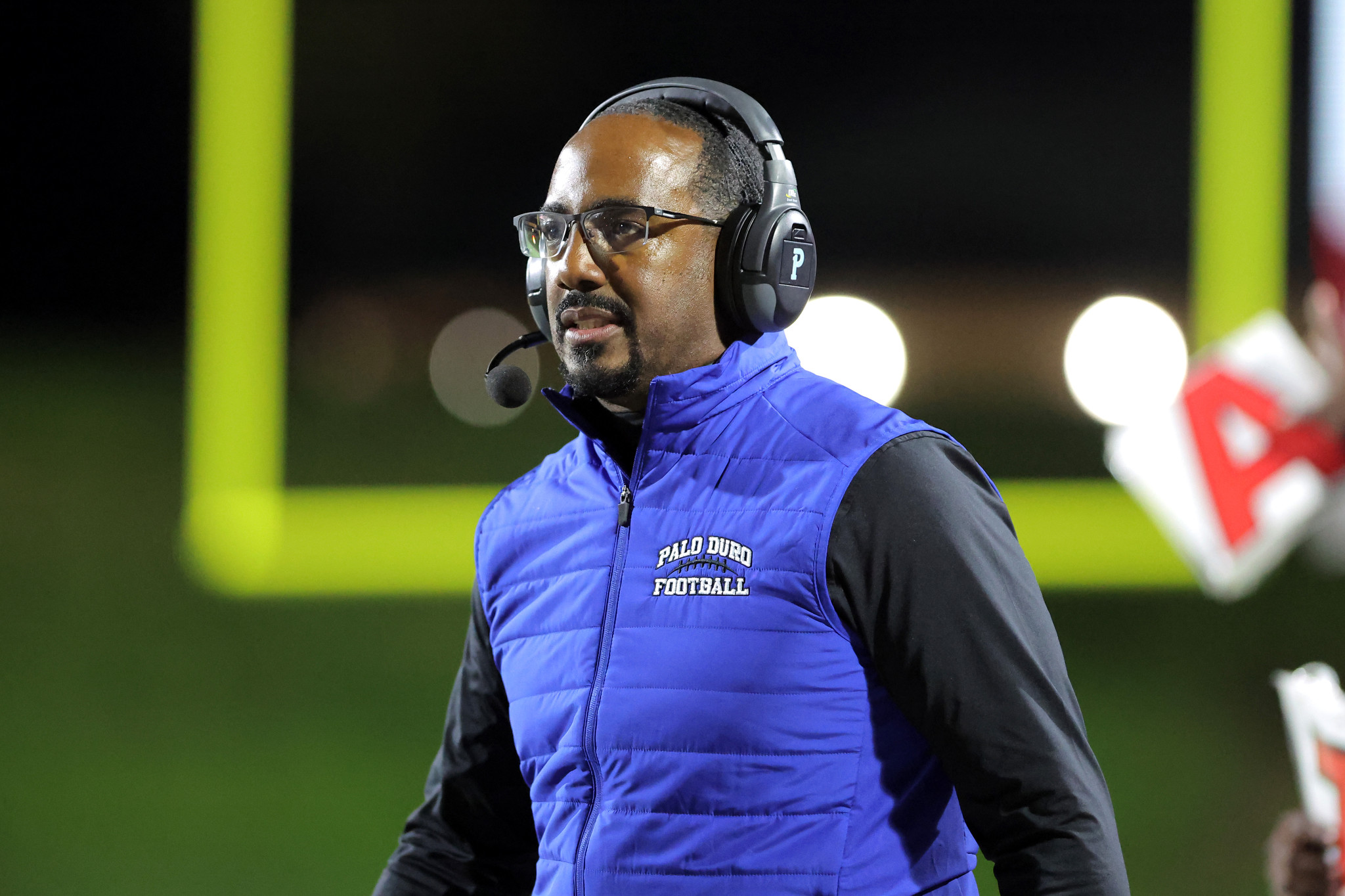 Coach wearing a blue padded vest and headset, speaking on a football field at night.