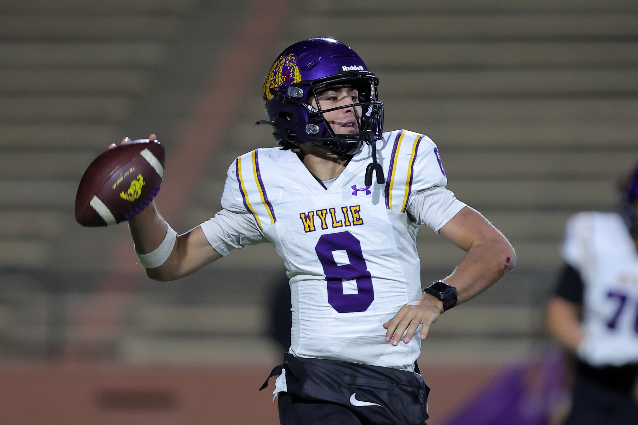 Quarterback in a white and purple Wylie jersey (number 6) throwing a football during a game, purple helmet on.