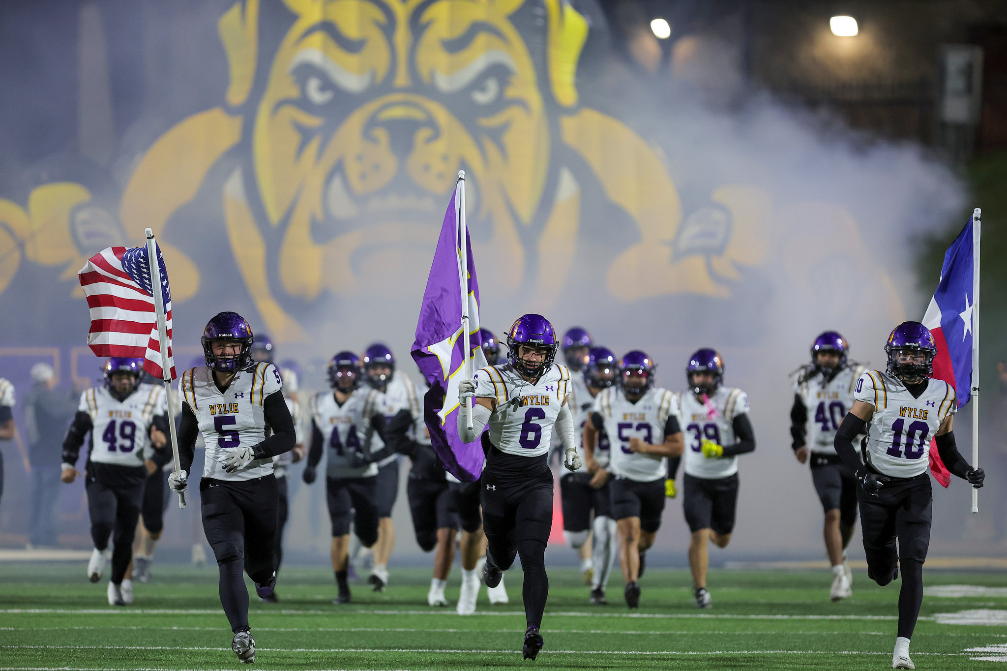 College football team in white and purple uniforms runs onto the field through smoke, carrying purple and white flags with a large bulldog mural in the background.