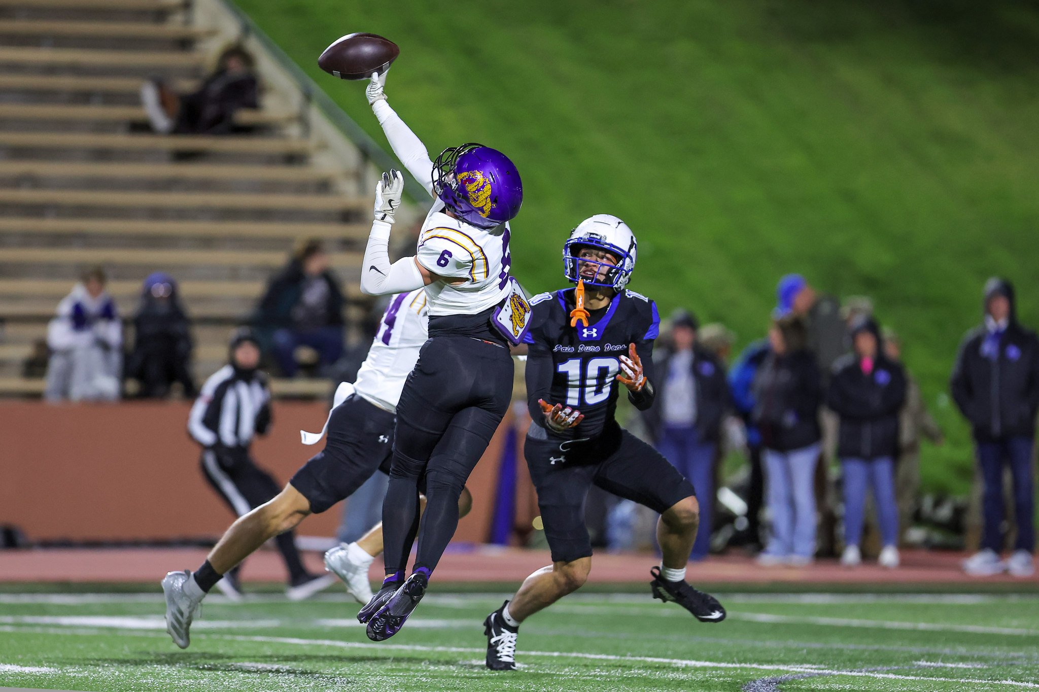 Football players leap for a catch: white-purple jersey in foreground reaching for a brown football while a defender in blue watches.
