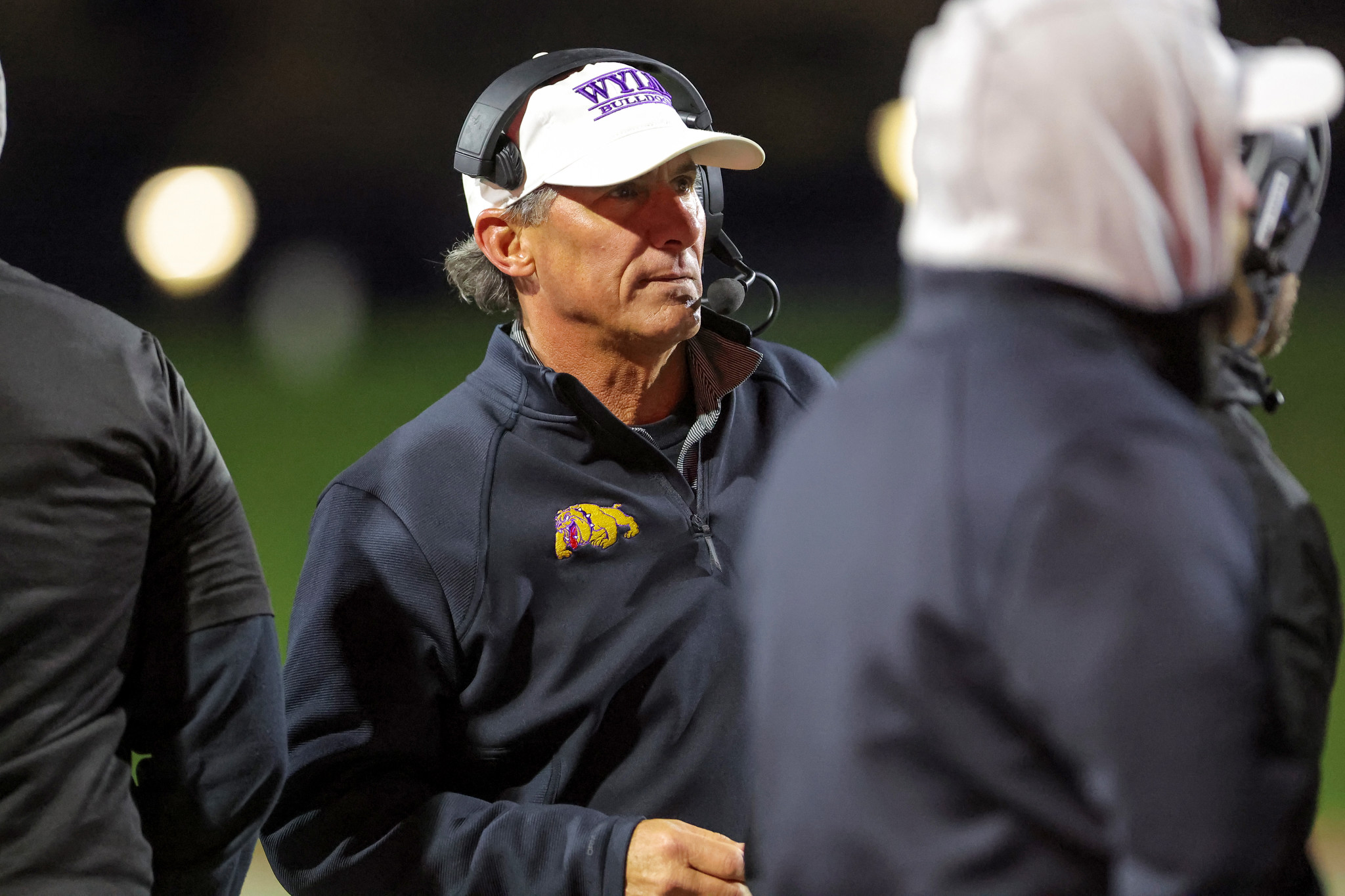 Coach with headset and white cap featuring a purple bulldog logo on a nighttime football field