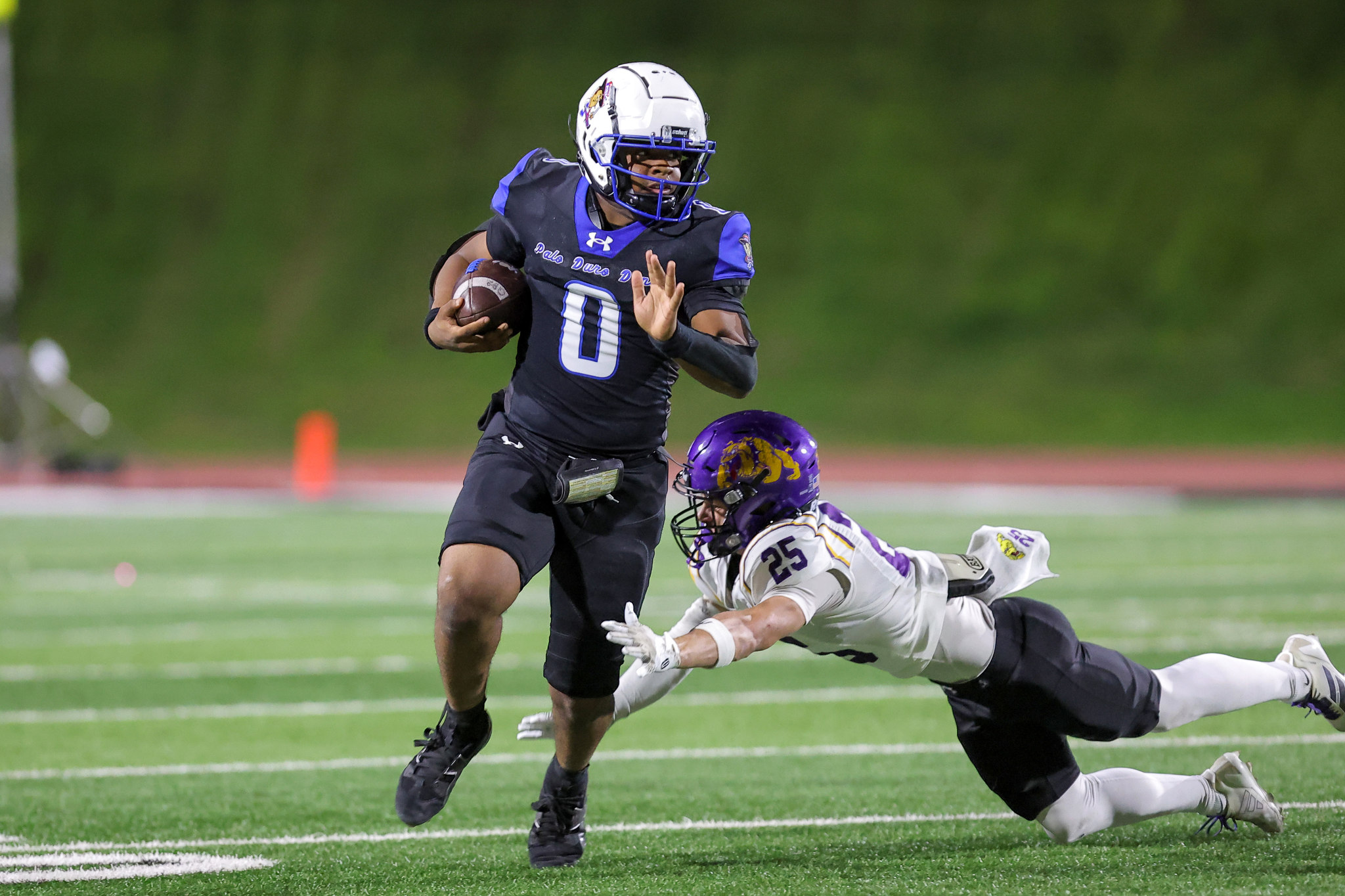 A football player in a black and blue uniform runs with the ball while a defender dives at his legs on a green field.