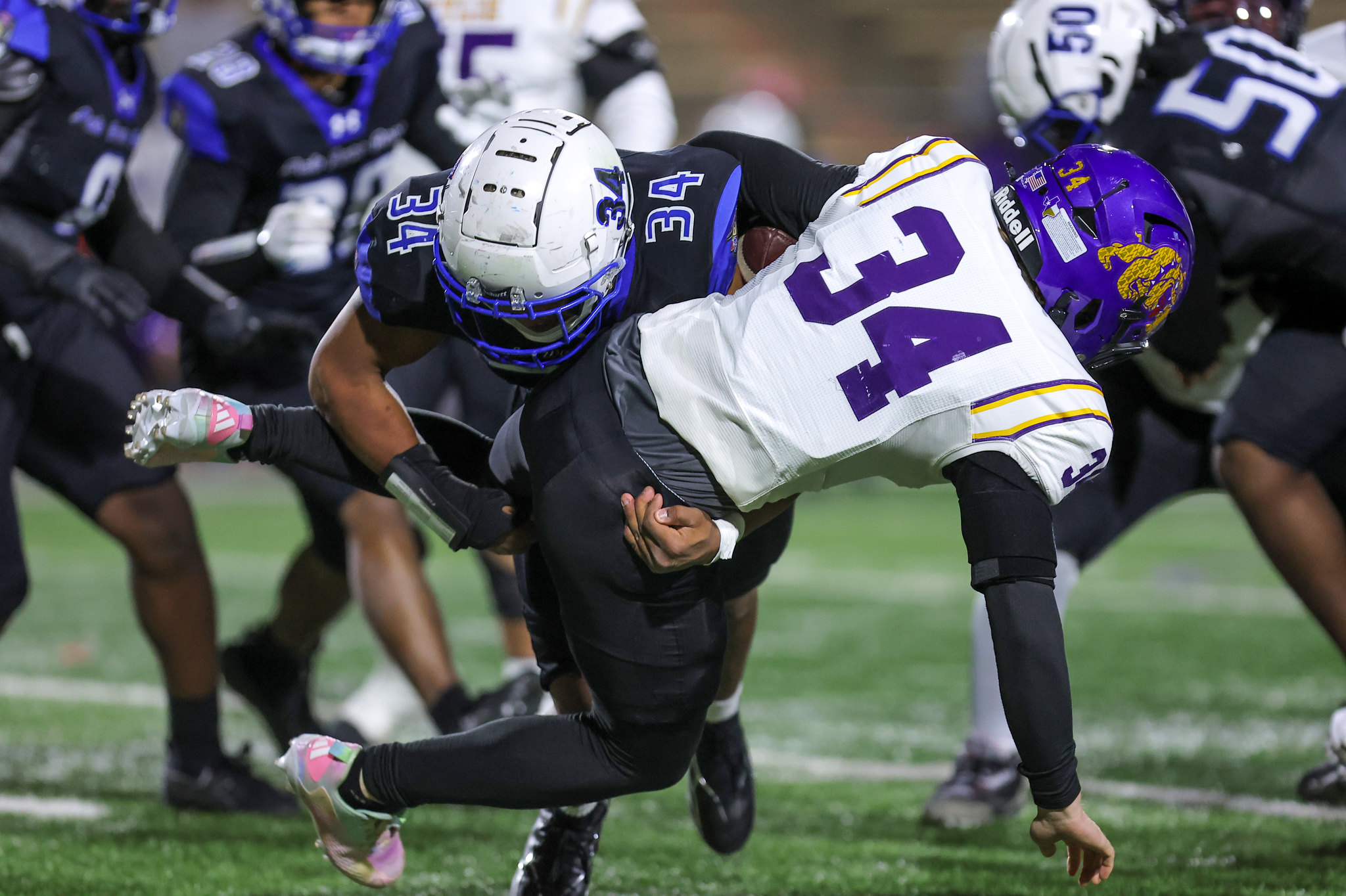 Two football players collide in a tackle on a grassy field, one in a white and purple jersey (#11) and the other in a dark uniform with a white helmet, surrounded by teammates and the goal line in the background.