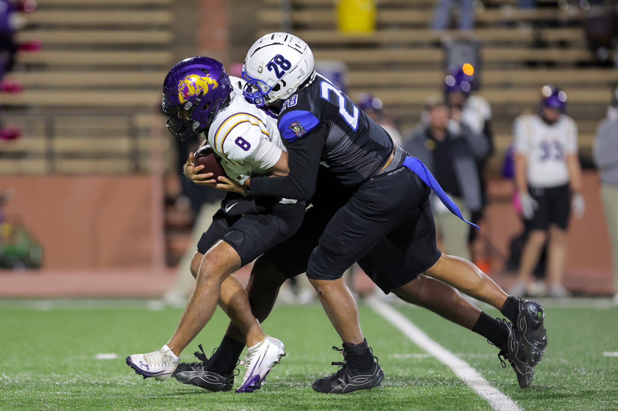 Two football players collide as they fight for the ball on a green turf field; one in a white jersey with purple helmet, the other in a dark uniform with a white helmet labeled 28.