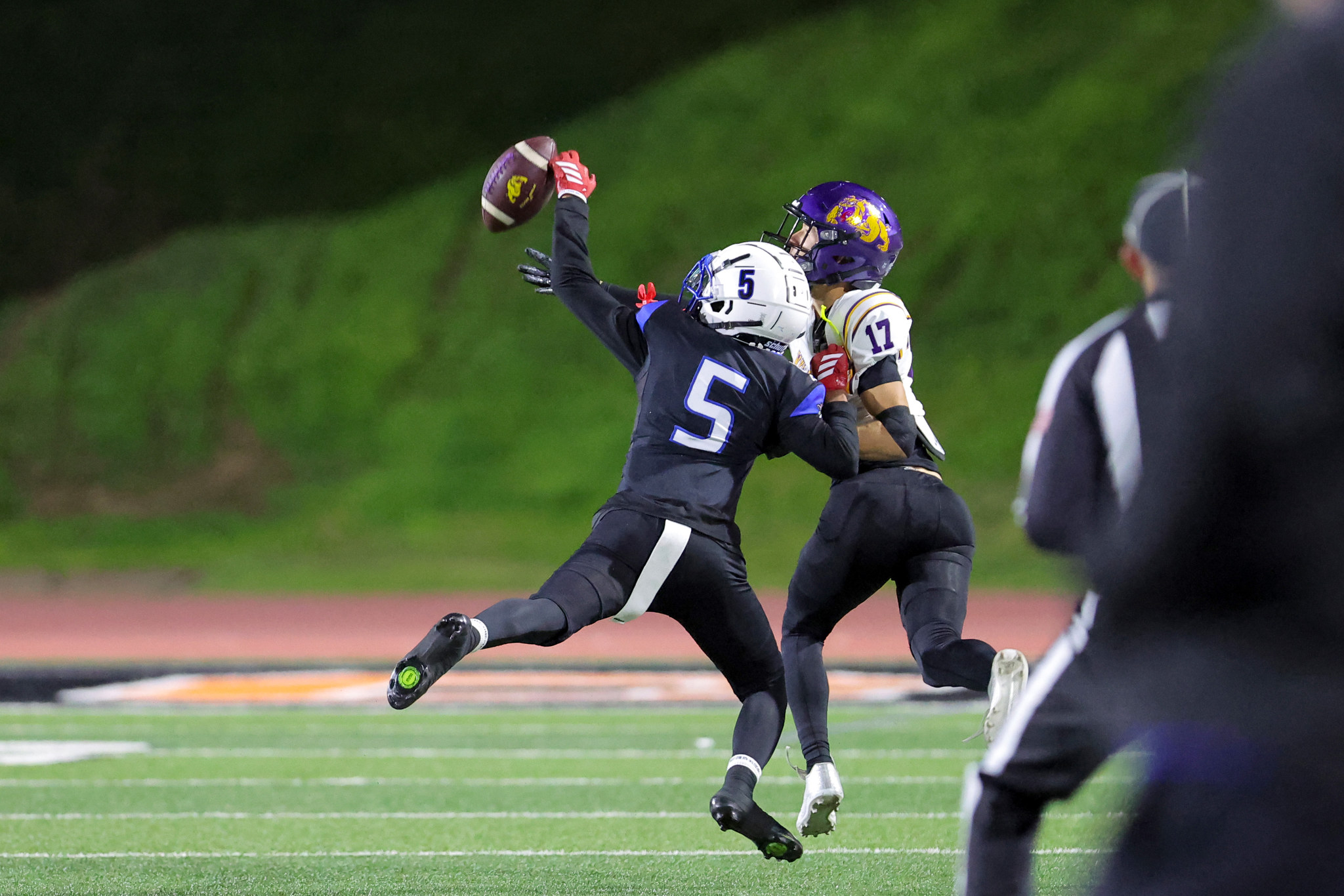 Two football players fight for a loose ball in midair; one in a white helmet reaches up while a purple-helmet opponent closes in.