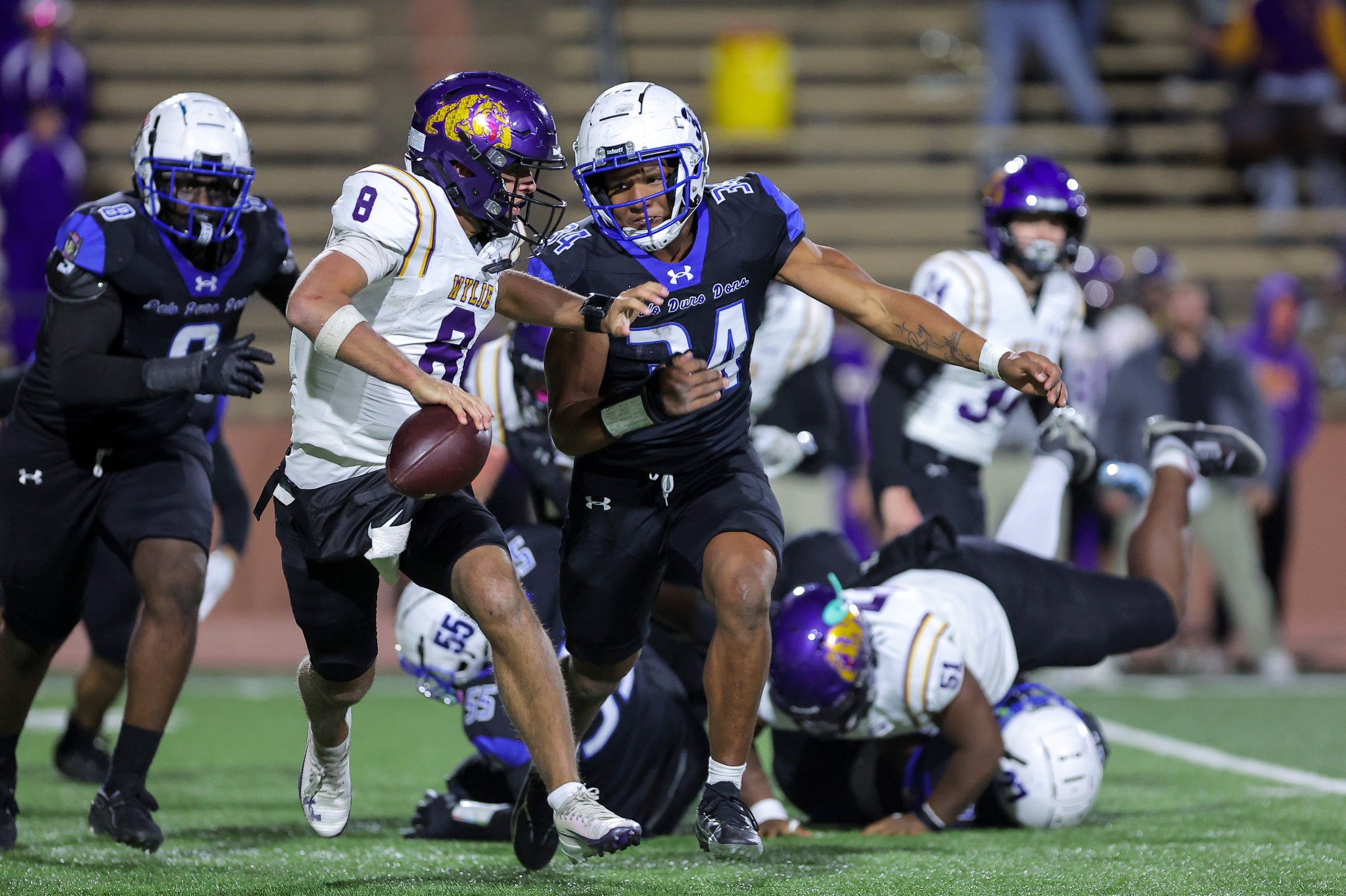 Two football players collide during a play, one in a white jersey (#8) and the other in a dark purple/blue jersey (#34), racing for the ball on a green turf field with teammates and spectators in the background.