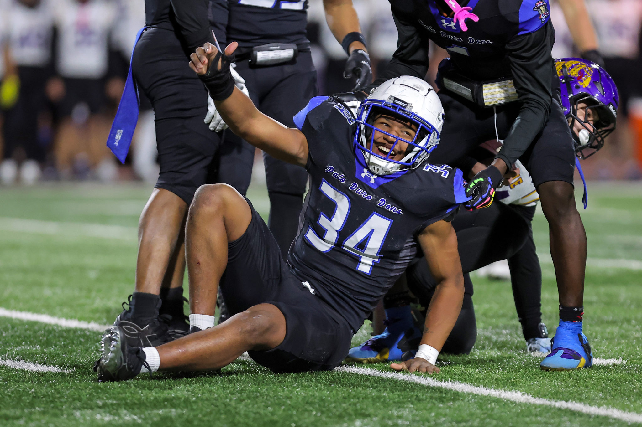 Football players in black uniforms tackle and fall on a green turf, central player on the ground smiling with a blue faceguard helmet nearby.