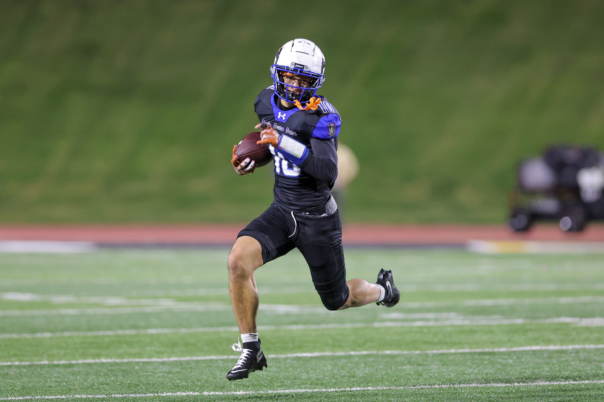 American football player in a dark uniform and white helmet sprinting with the ball on a green field.
