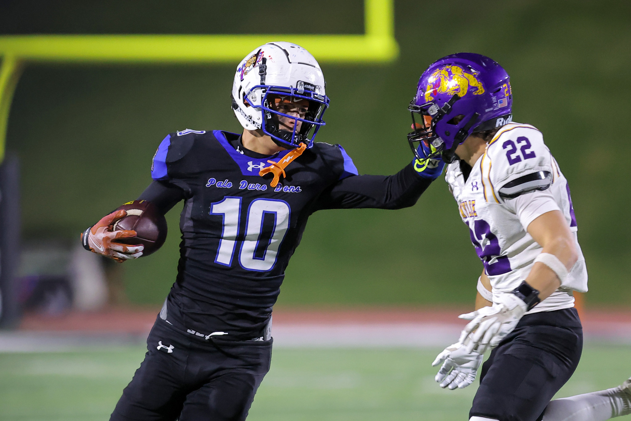 Two football players collide/challenge for the ball on a field during a night game; one in a black uniform with number 10 and white helmet, the other in white with purple accents and a purple helmet.
