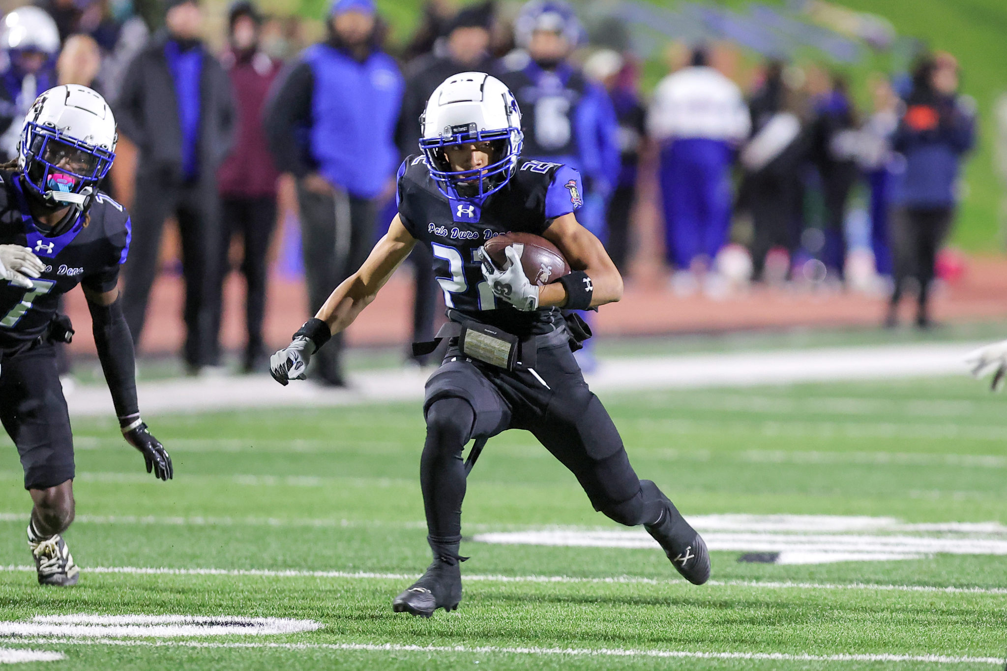 A football player in blue-black uniform carries a ball while sprinting down the field, defenders behind him on a stadium track.