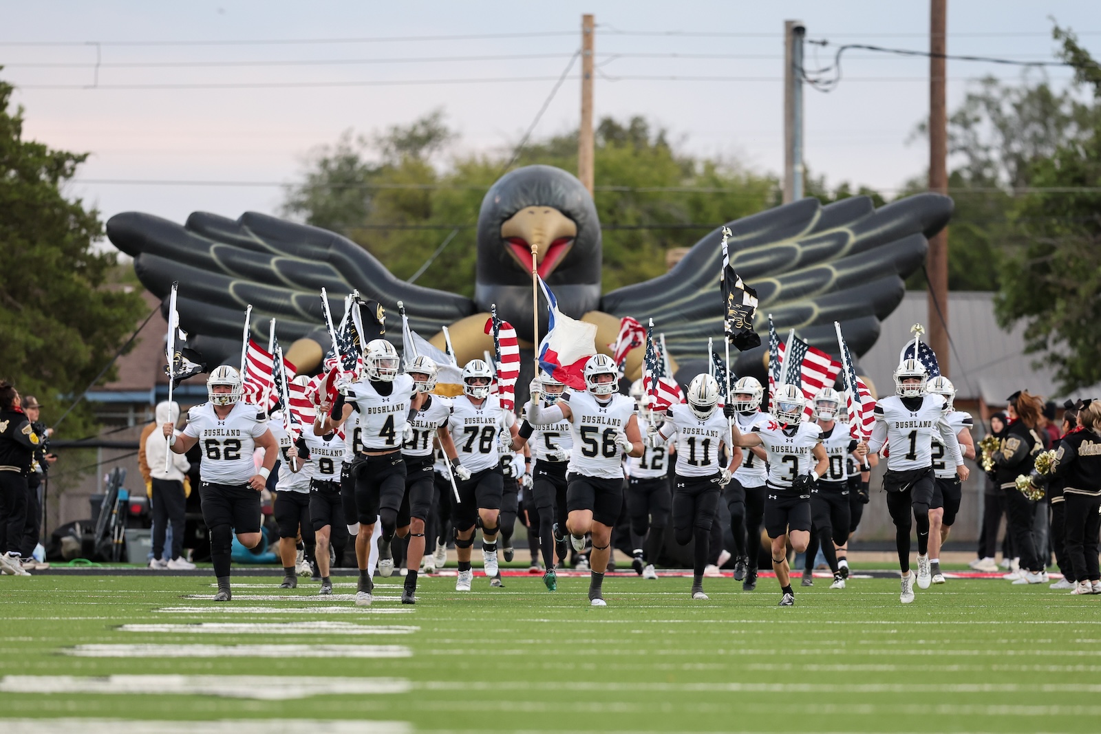 Seabolt Chase-15981 | Press Pass Sports Bushland high school football players run onto the field waving American flags, with a large inflatable turkey in the background.