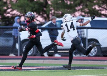 Seabolt Chase-16315 | Press Pass Sports Two football players sprint on a track field, one in a black and red uniform chasing the ball, the other in a white jersey number 14 pursuing him.