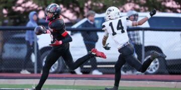 Two football players sprint on a track field, one in a black and red uniform chasing the ball, the other in a white jersey number 14 pursuing him.