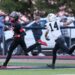 Seabolt Chase-16315 | Press Pass Sports Two football players sprint on a track field, one in a black and red uniform chasing the ball, the other in a white jersey number 14 pursuing him.