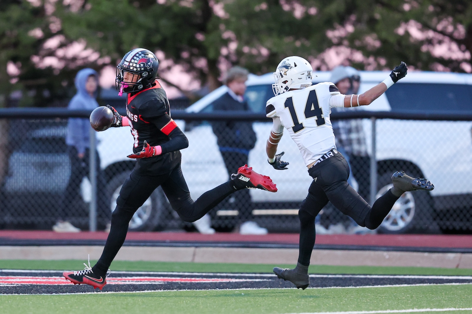 Seabolt Chase-16315 | Press Pass Sports Two football players sprint on a track field, one in a black and red uniform chasing the ball, the other in a white jersey number 14 pursuing him.