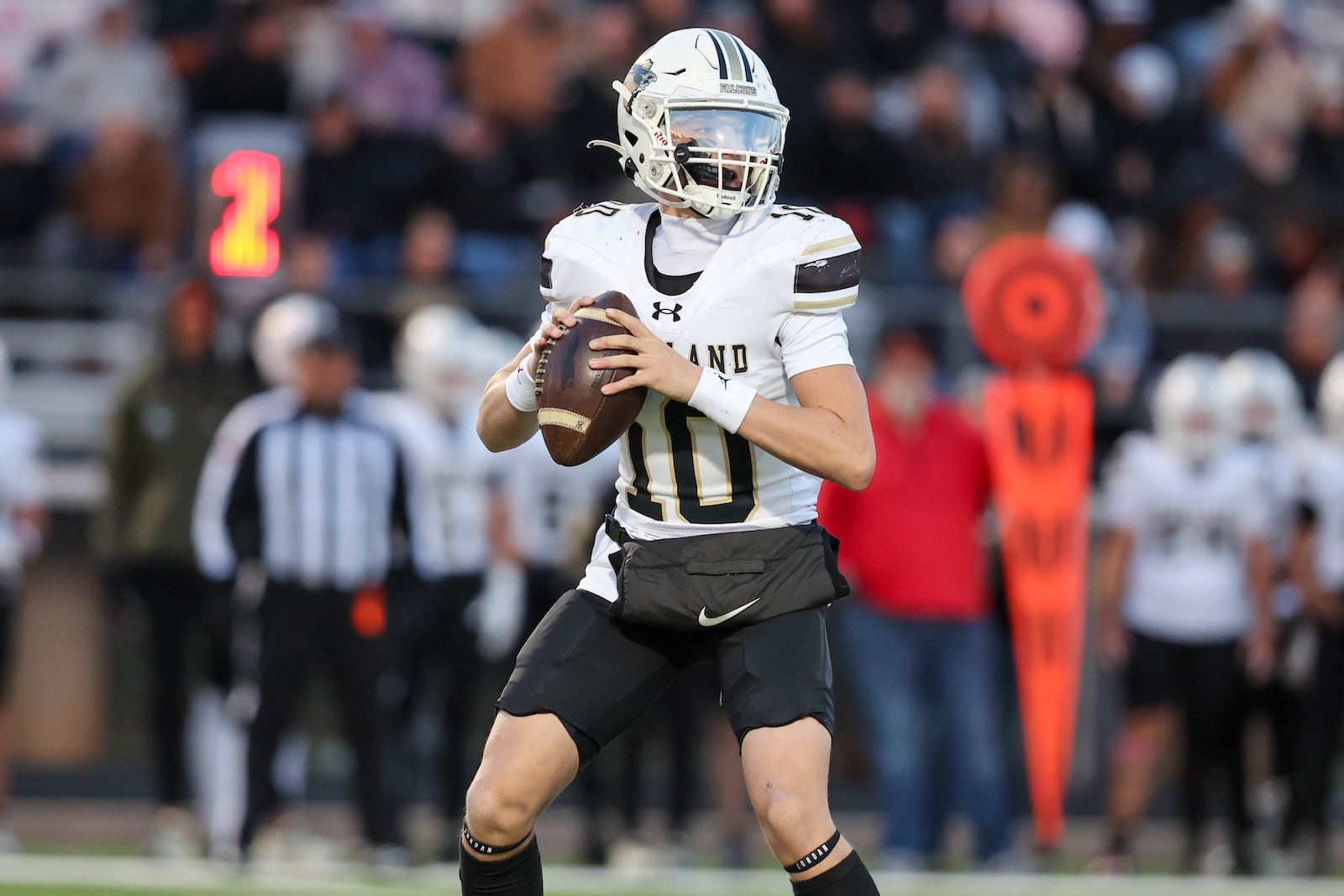 Seabolt Chase-16442 | Press Pass Sports Quarterback in white and black uniform holds football, preparing to throw on a football field with fans in the stands behind him.