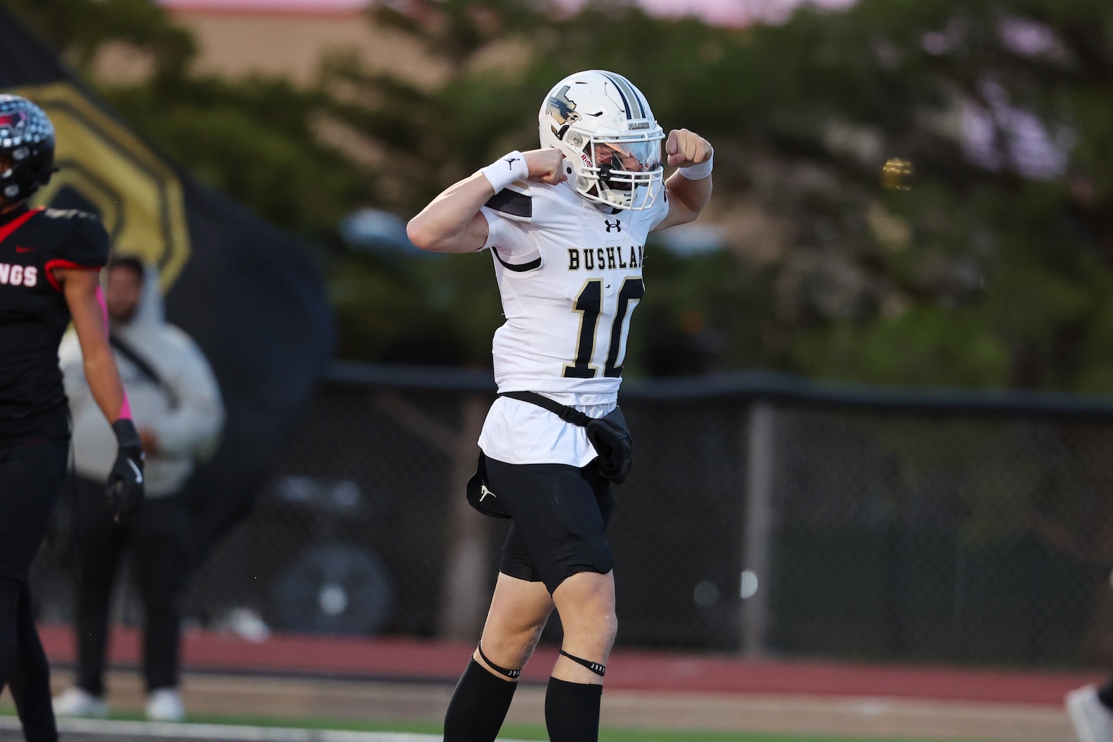 Seabolt Chase-16591 | Press Pass Sports Football player in a white Bushland jersey (number 10) flexing arms in celebration on a football field, with a defender in black nearby.