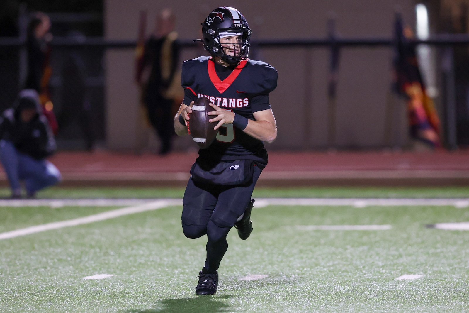Seabolt Chase-17239 | Press Pass Sports Football player in navy and red Mustangs uniform runs with the ball on a lit turf field at night."Mustangs" logo on helmet visible.