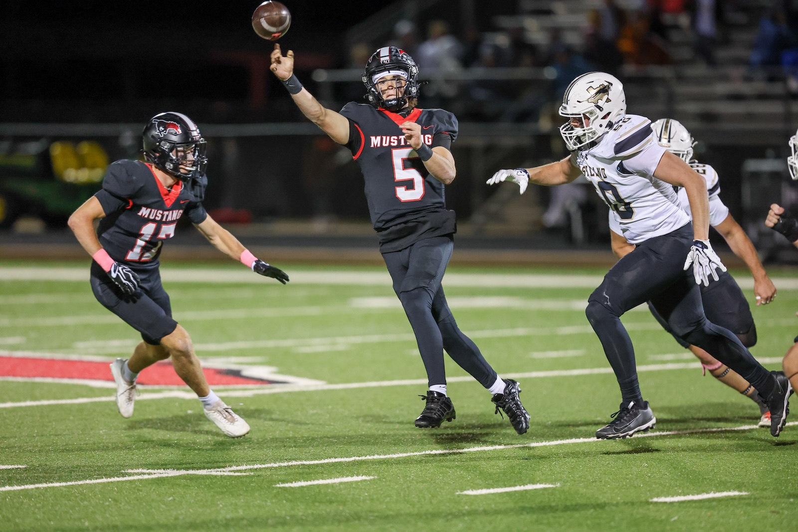 Seabolt Chase-17334 | Press Pass Sports Football game at night: quarterback (number 5) in a black Mustang jersey throws a pass while teammates and defenders chase on a bright green field.