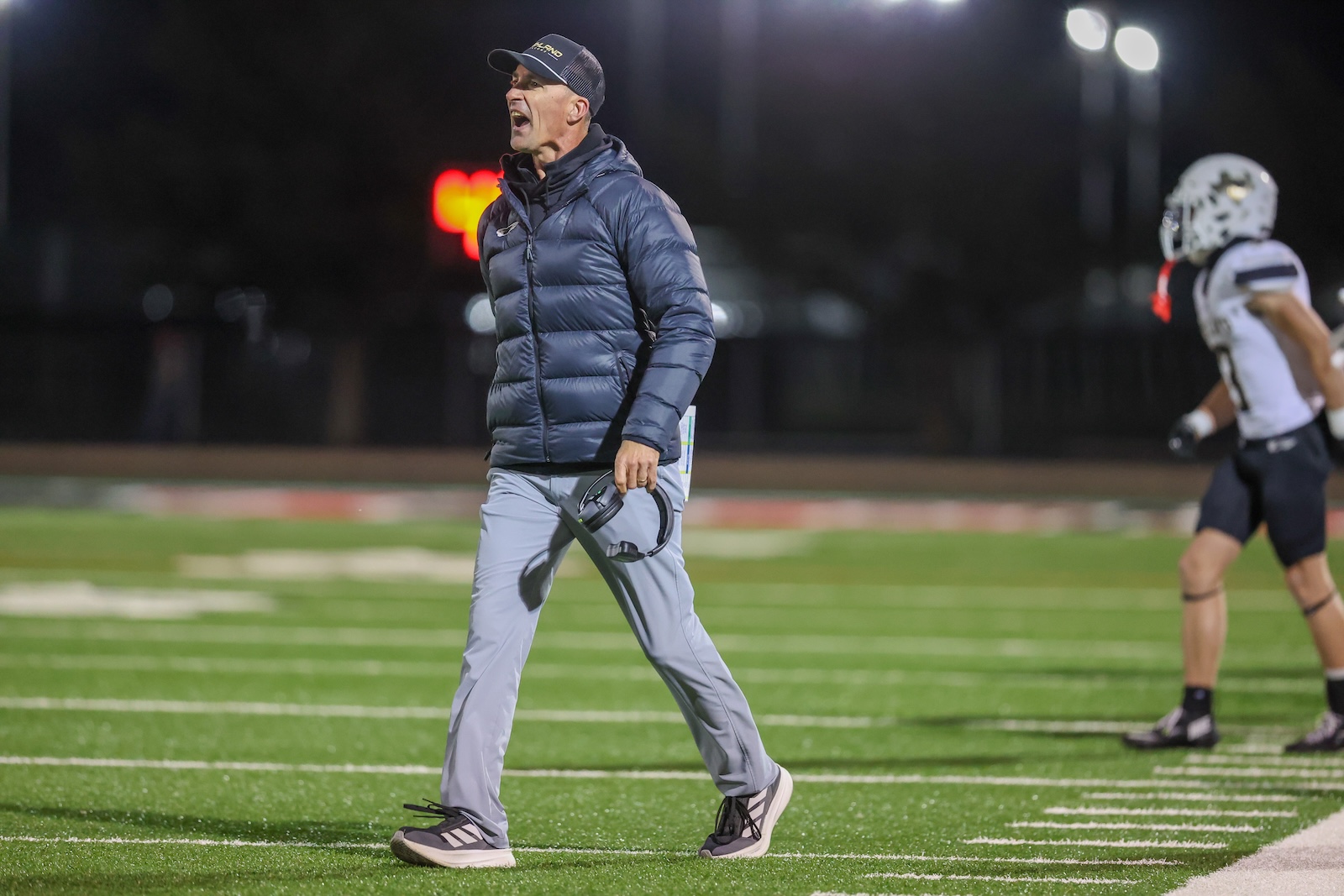 Seabolt Chase-17391 | Press Pass Sports Coach in a blue puffer jacket shouting on a lit football field at night, headset in hand.