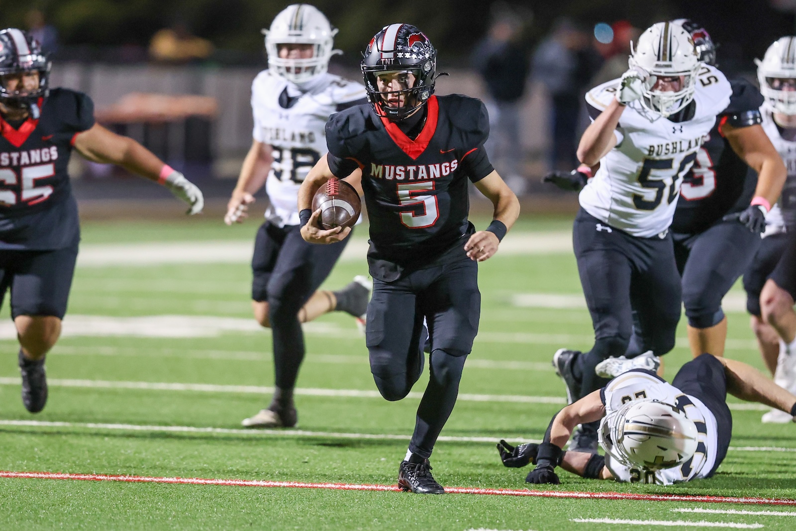 Seabolt Chase-17471 | Press Pass Sports Football player (Mustangs) wearing number 5 runs with the ball as defenders close in on a night game on a sunny field edge.