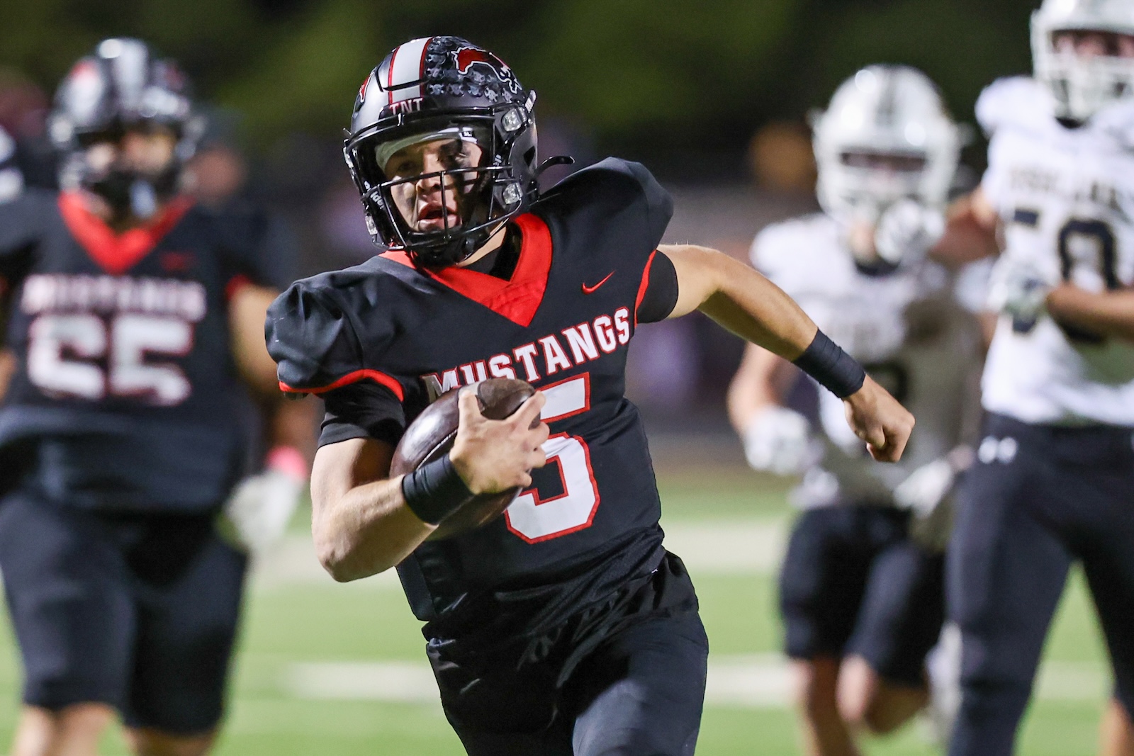 Seabolt Chase-17489 | Press Pass Sports Football player in a black Mustang uniform runs with the ball during a game on a green field.