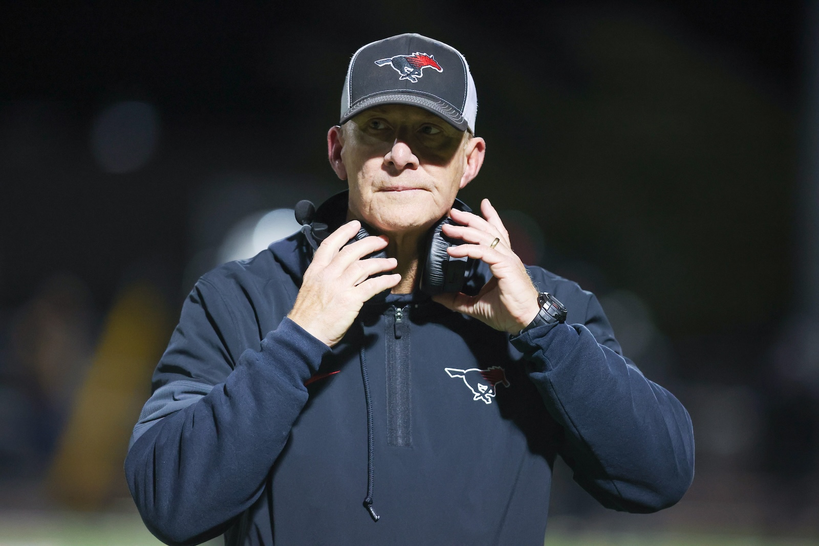 Seabolt Chase-17647 | Press Pass Sports Coach in a navy jacket and cap with a horse logo, adjusting a wireless headset on a football field at night.
