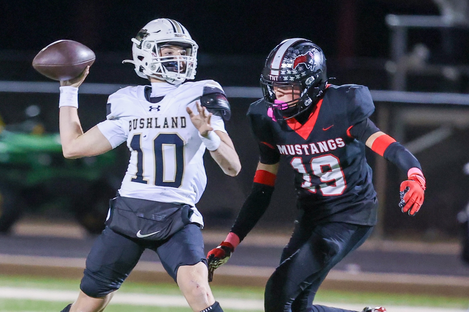Seabolt Chase-17668 | Press Pass Sports Bushland quarterback #10 in white throws a pass as a Mustang defender in black (#39) pressures him during a night game.
