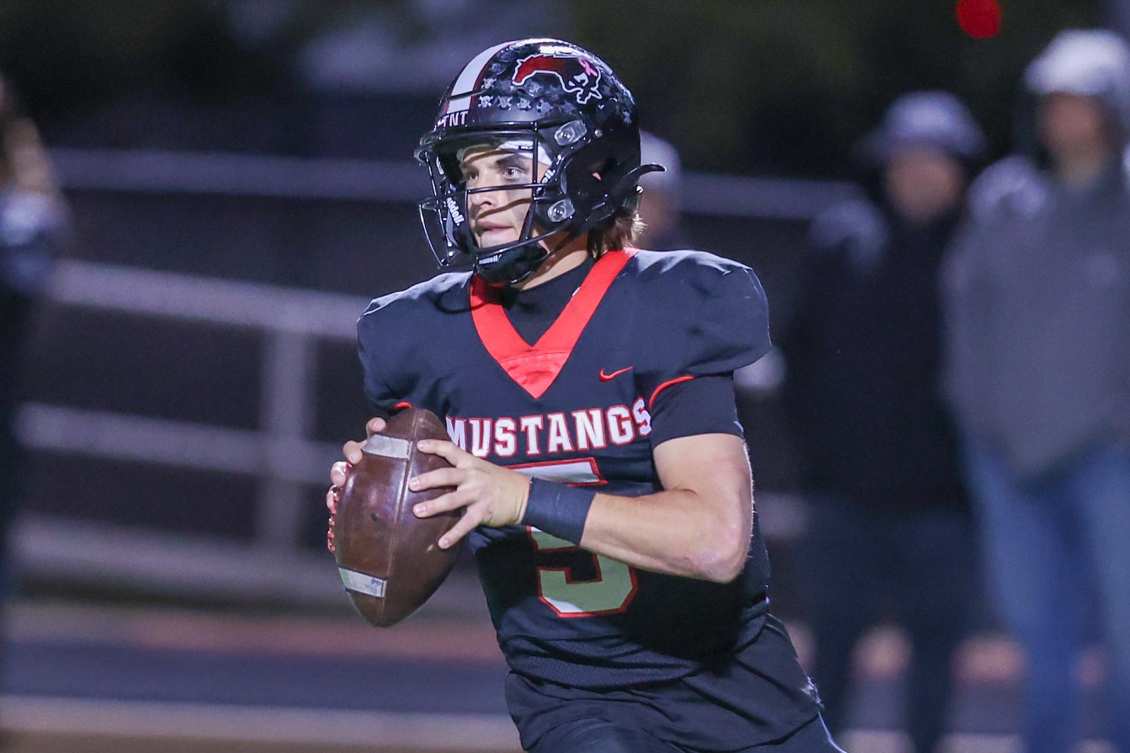 Seabolt Chase-17677 | Press Pass Sports Football quarterback in dark helmet and red-trimmed uniform holding a ball, running on field for the Mustangs