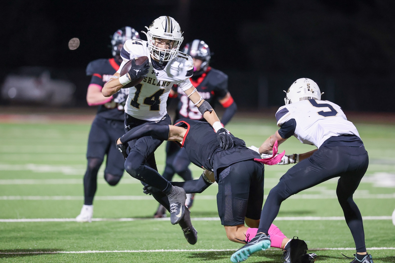 Seabolt Chase-17732 | Press Pass Sports A football player in a white jersey #14 runs with the ball while being chased by opponents on a night game under stadium lights, on a green artificial turf field.