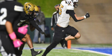 Football players sprinting on the field during a game, white jersey with number 25 leading the run behind a black-clad opponent in gold helmet