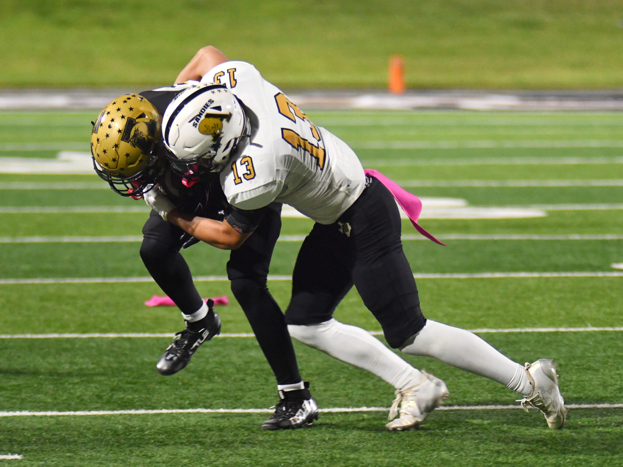 DSC_5536 | Press Pass Sports Two football players tackle each other on a green turf, wearing white and gold uniforms and helmets.