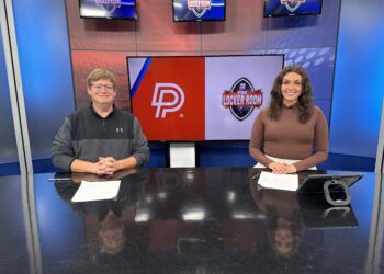 IMG_8345 | Press Pass Sports Two news anchors sit at a dark desk in a blue TV studio with screens behind them showing 'The Locker Room' logo and a red-white graphic.