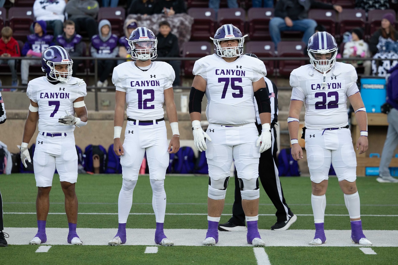 CHS-WPHS-FB-4 | Press Pass Sports Four Canyon football players in white uniforms stand on the field, numbers 7, 12, 75, and 22 visible, purple socks and helmets.