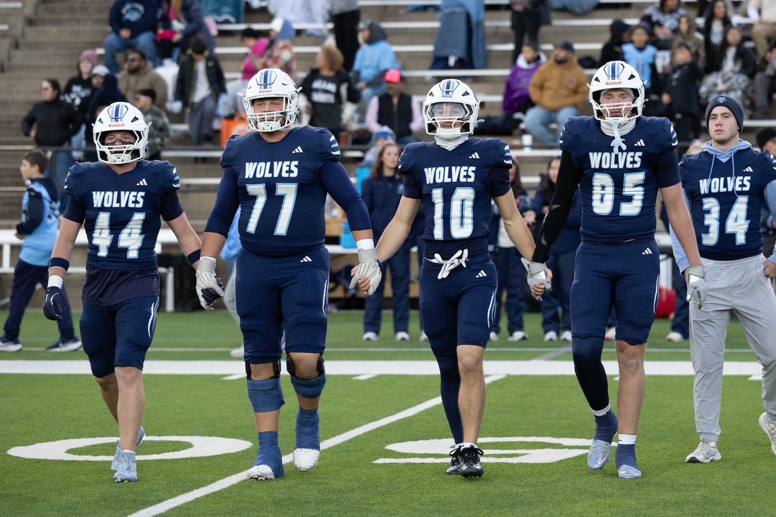 CHS-WPHS-FB-5 | Press Pass Sports Five Wolves football players in blue uniforms walk hand in hand across a green field with a crowded stadium behind them.